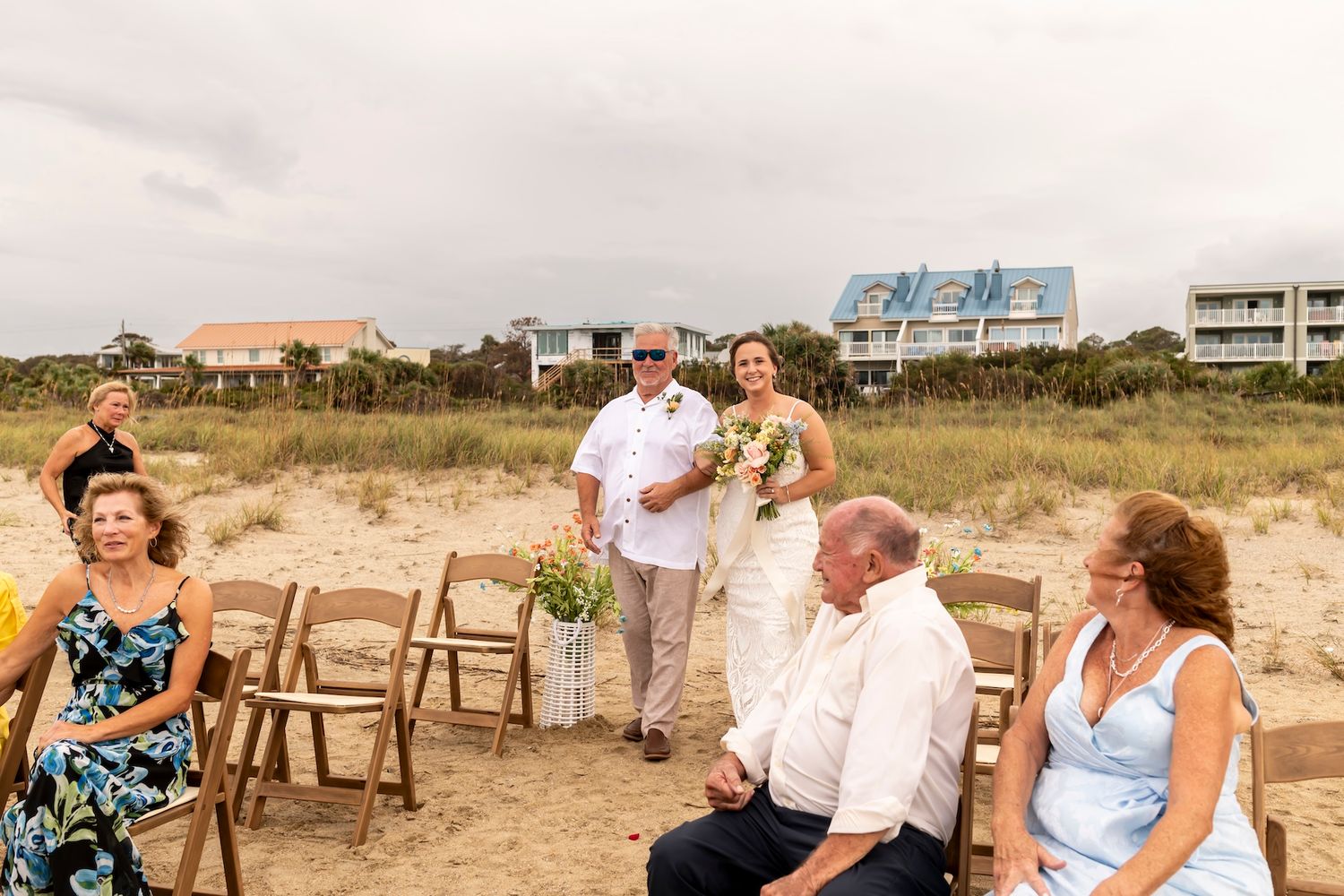 Wedding ceremony guests seated in wooden chairs on a beach with beach houses and buildings visible in the background.