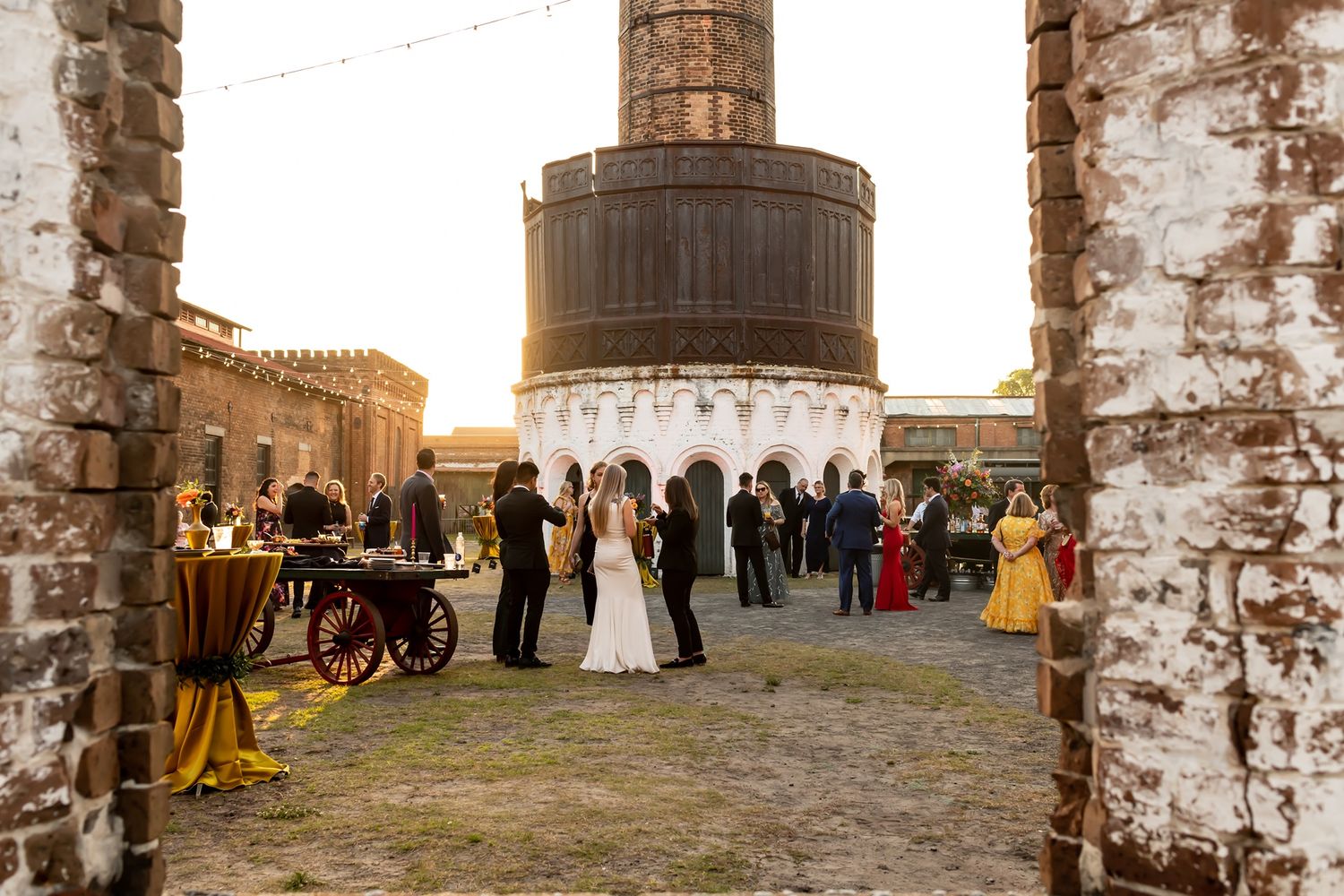 A wedding celebration at sunset in a rustic stone courtyard with guests gathered around an industrial-style tower structure.