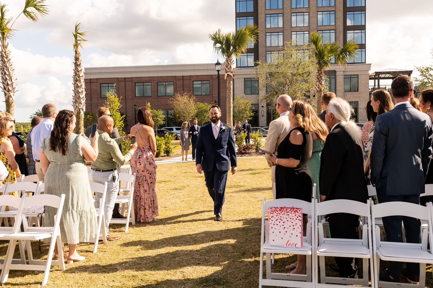 A groom walks down an outdoor aisle at a waterfront wedding ceremony with palm trees and white chairs setup for guests.
