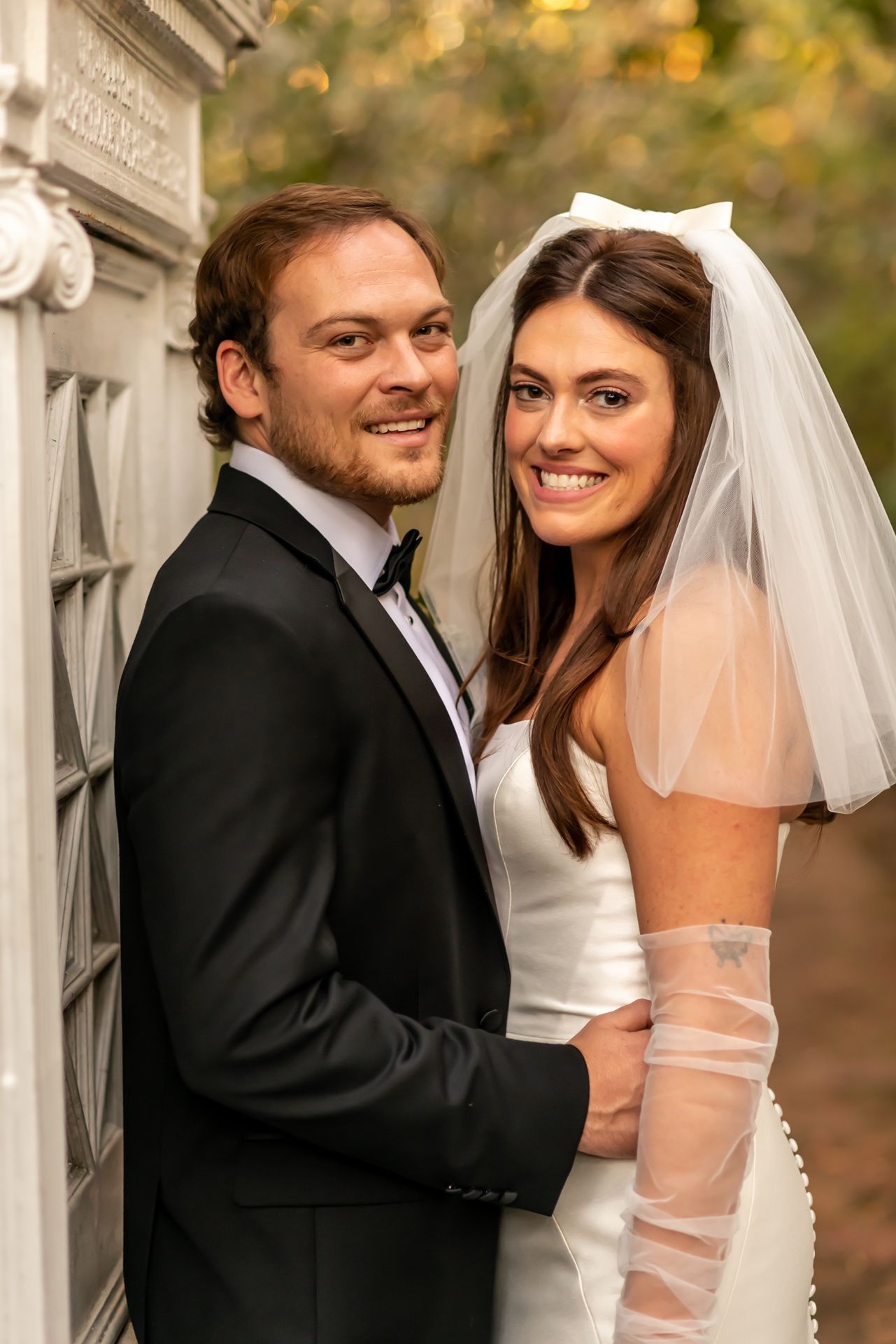A couple in wedding attire share romantic moments against a light-filled outdoor backdrop near a stone pillar.