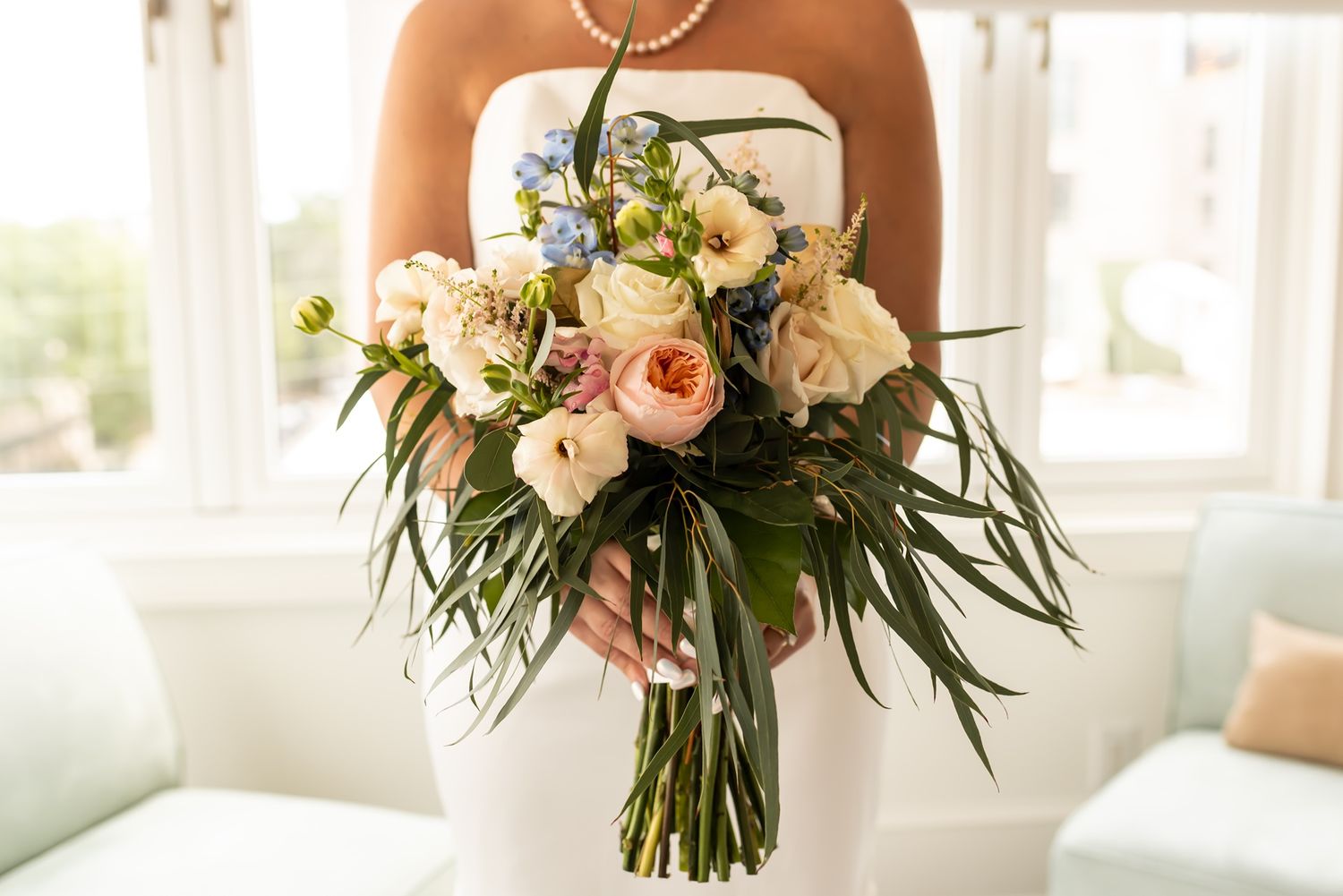 A delicate pink and white floral bouquet with roses and greenery sits by a sunlit window.