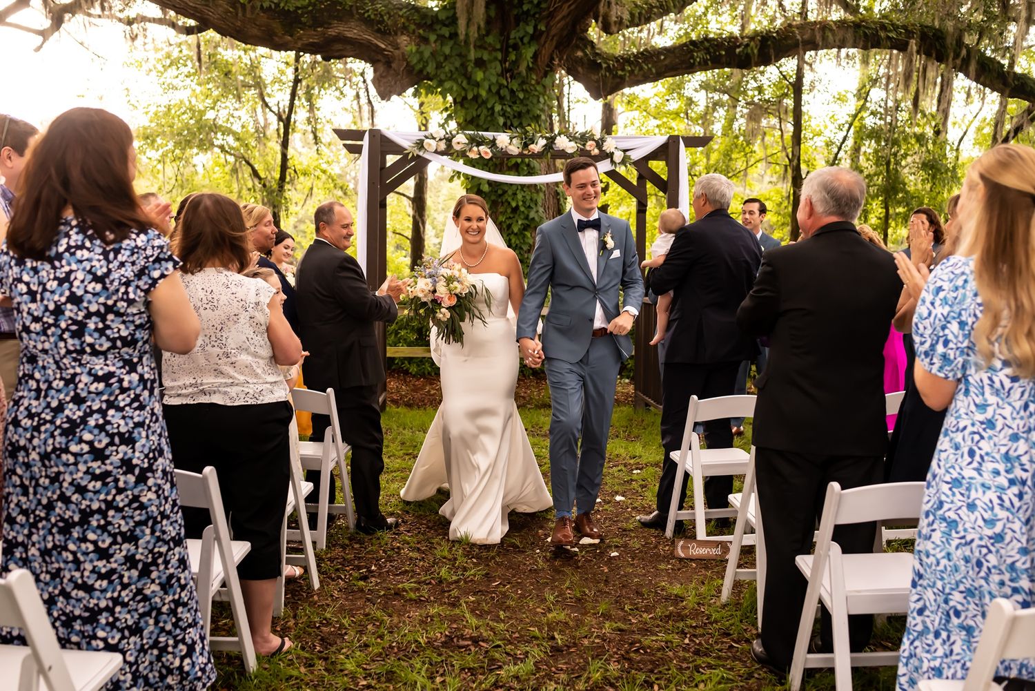 Newlyweds walk down the aisle under a rustic garden archway as wedding guests in summer attire look on.