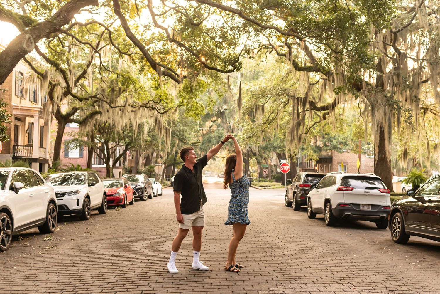 Pedestrians walking on an oak tree-lined cobblestone street in Savannah with Spanish moss overhead and parked cars along curb.