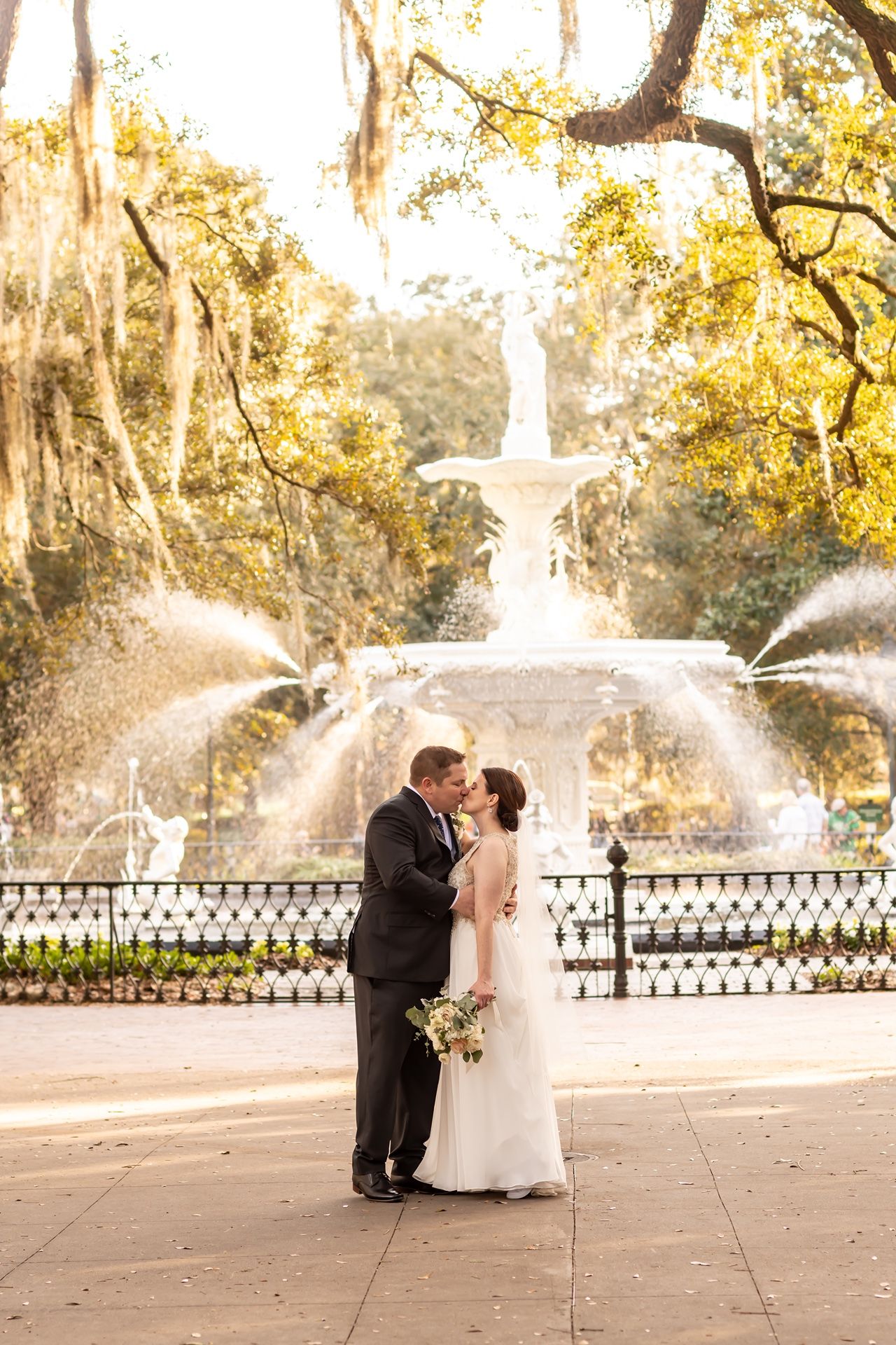 Golden sunlight streaming through trees as a couple shares a romantic moment by an ornate fountain in the park during sunset.