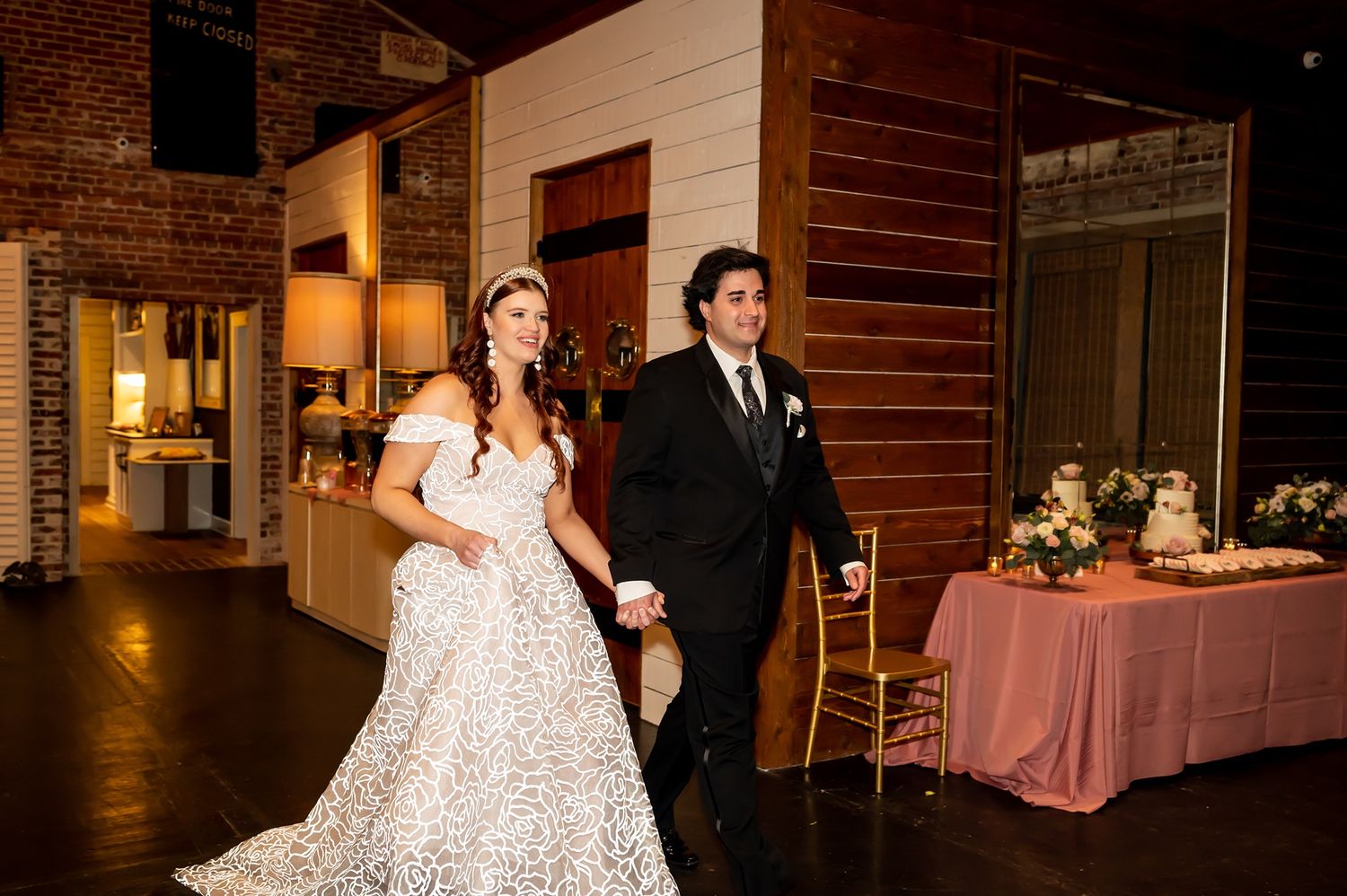 A pair in formal attire walk together in a rustic wood-paneled venue with warm lighting and elegant decor.