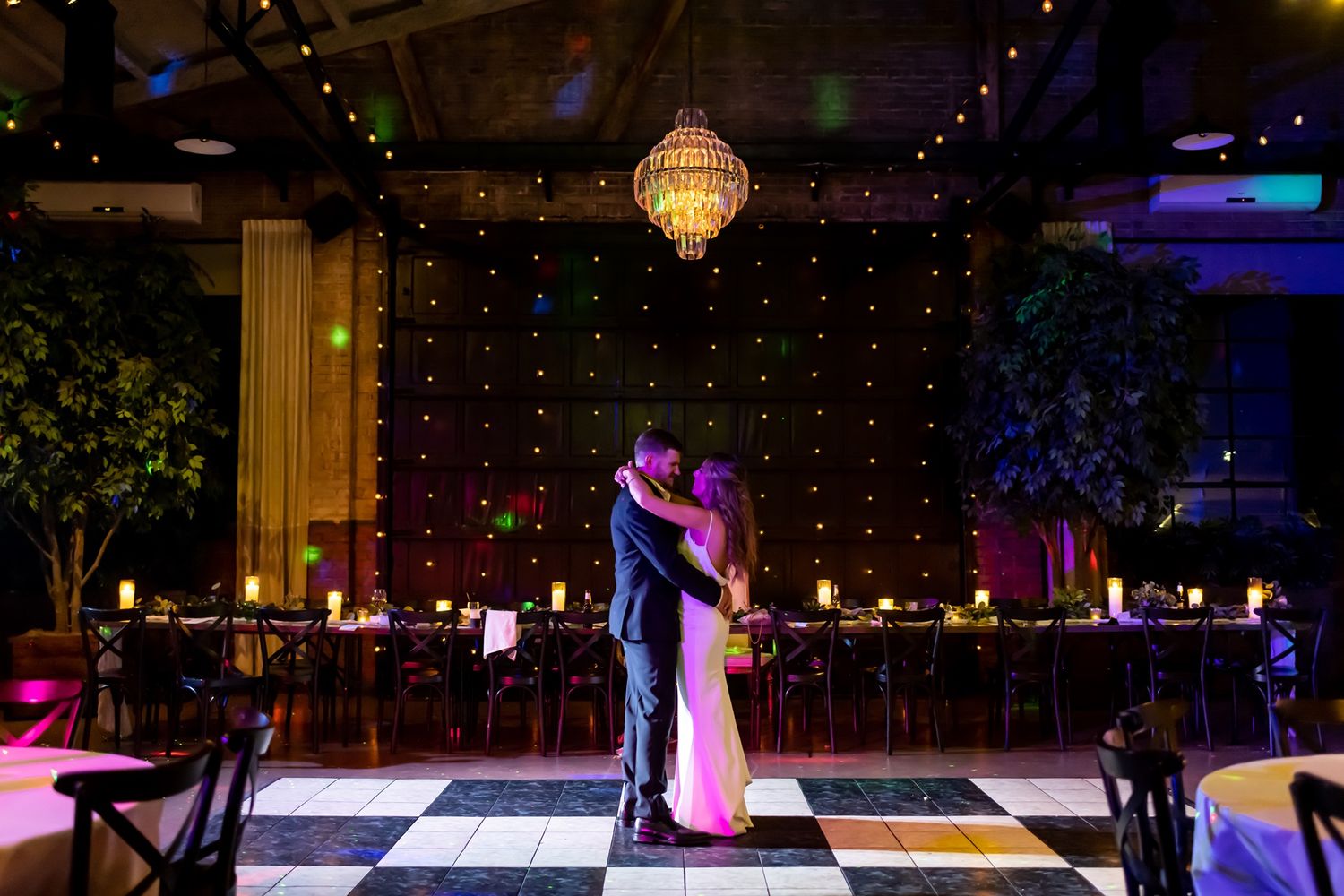 A romantic first dance at an evening wedding reception with chandelier and twinkle light backdrop on a black and white floor.