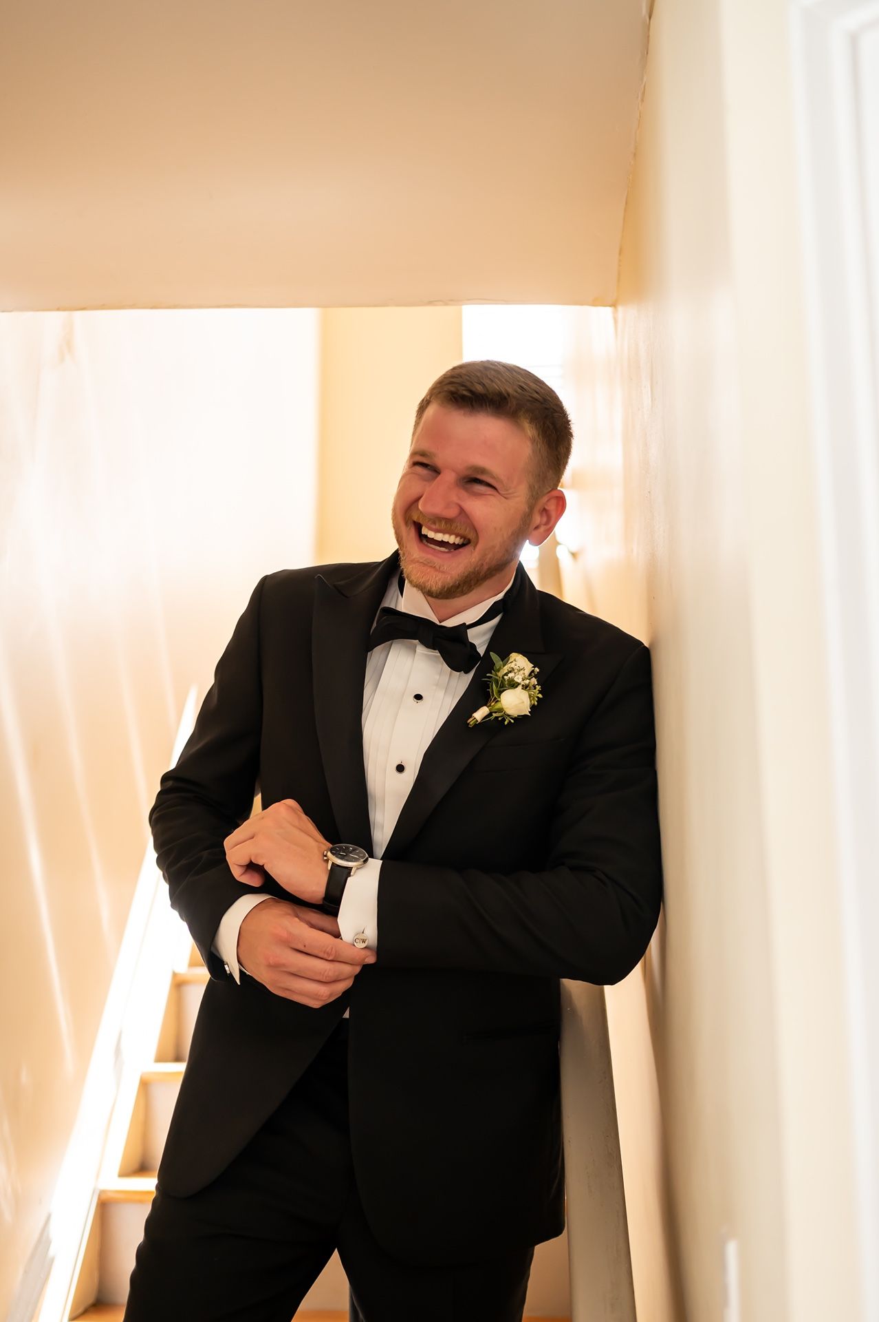 A groom in a classic black tuxedo laughs joyfully while adjusting his cuff links at a wedding.