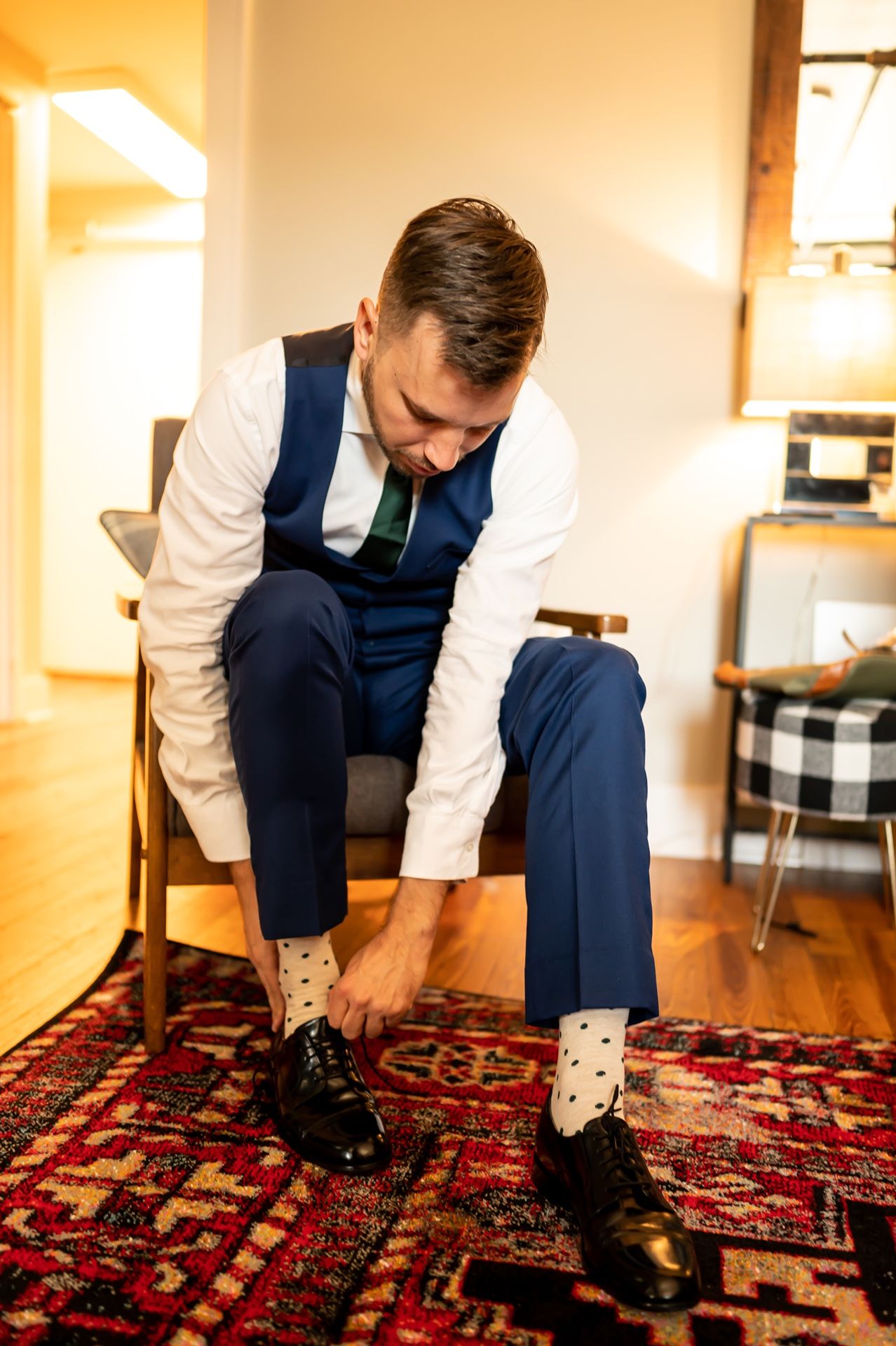 Person wearing a formal vest and dress pants ties their black dress shoes while sitting on a chair over a patterned rug.