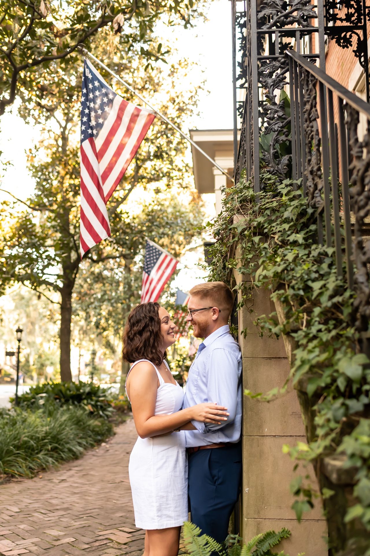A romantic couple embraces beneath American flags hanging in a garden walkway with brick pavers and lush greenery.