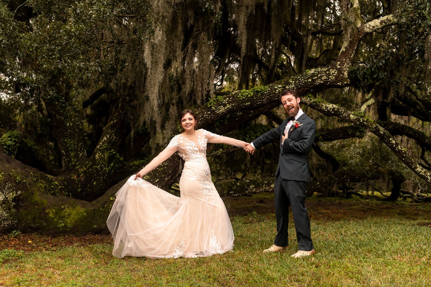 A couple in formal wedding attire poses playfully under moss-draped oak trees in a southern garden setting.