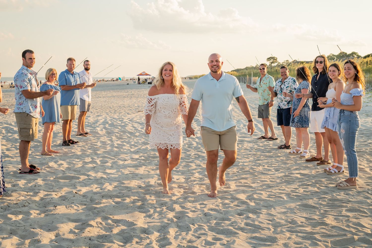 A couple walks along the beach surrounded by guests at sunset during a beach celebration event.