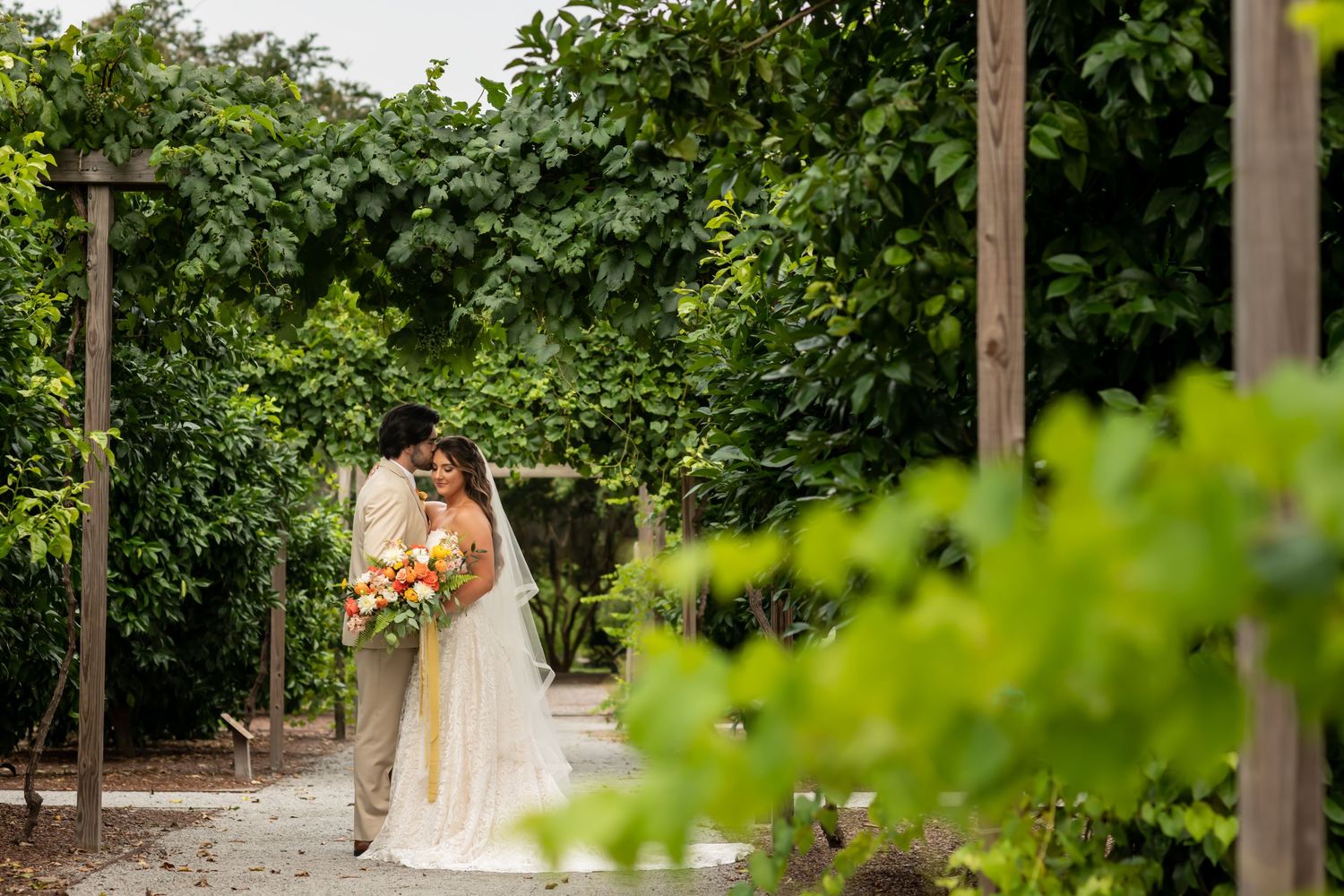 Two people share a romantic moment in a lush garden path lined with greenery and wooden posts.