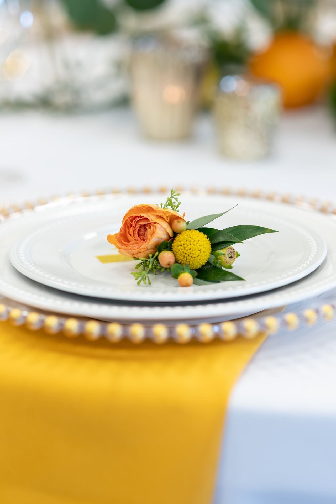 A delicate floral place setting with an orange ranunculus and yellow craspedia on a white plate with gold beaded charger.