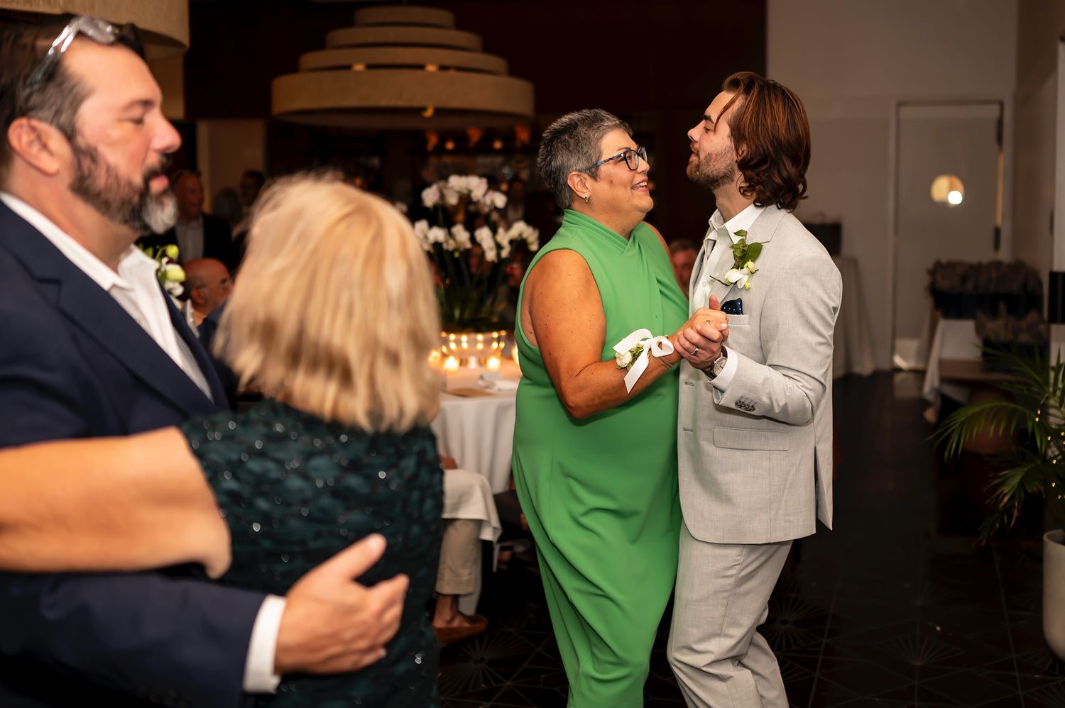 Guests dressed in green and formal attire mingle and dance together at a dimly lit social gathering or party.