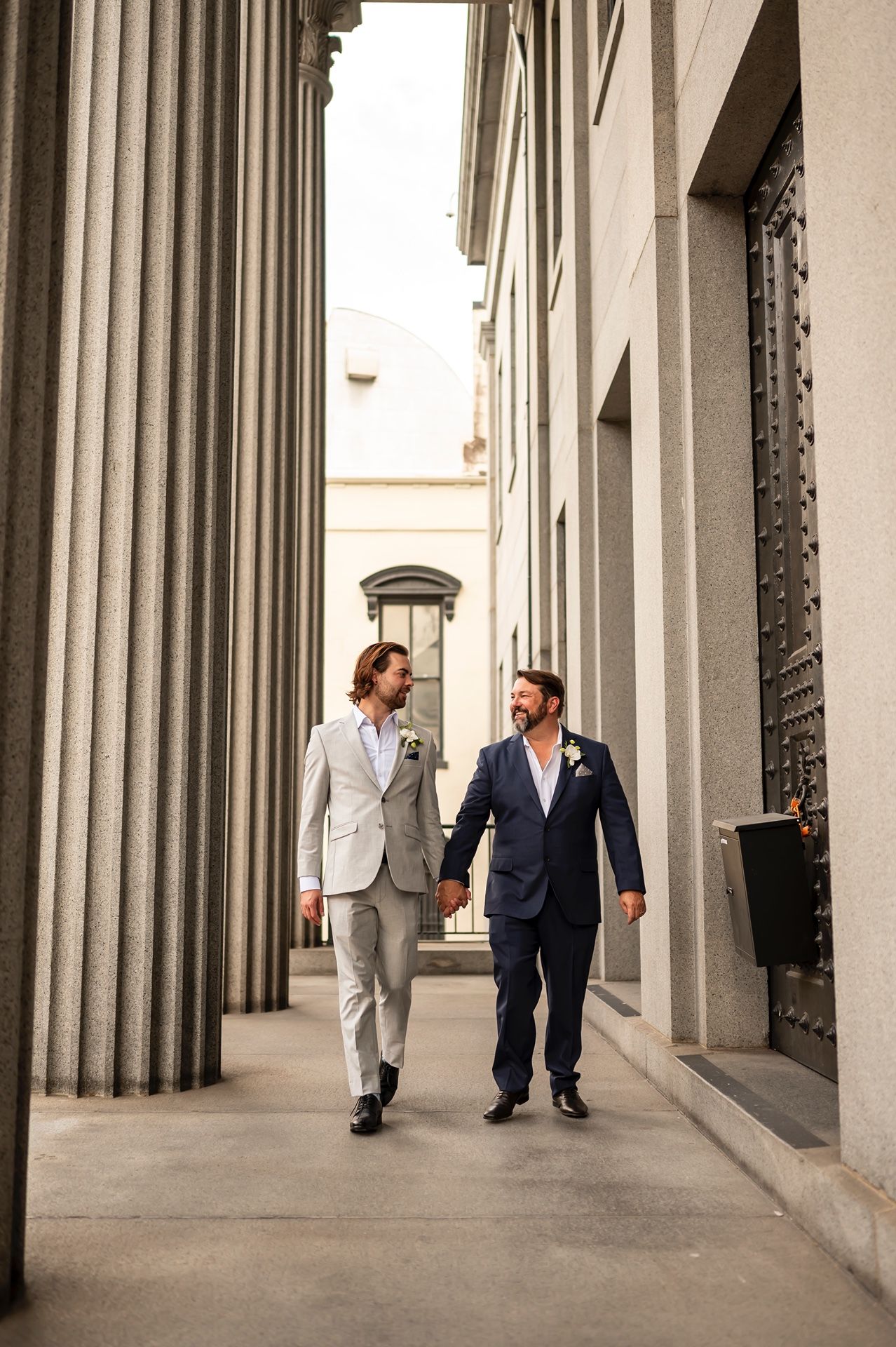 Two people in wedding attire walking together along a columned walkway outside a classic building.
