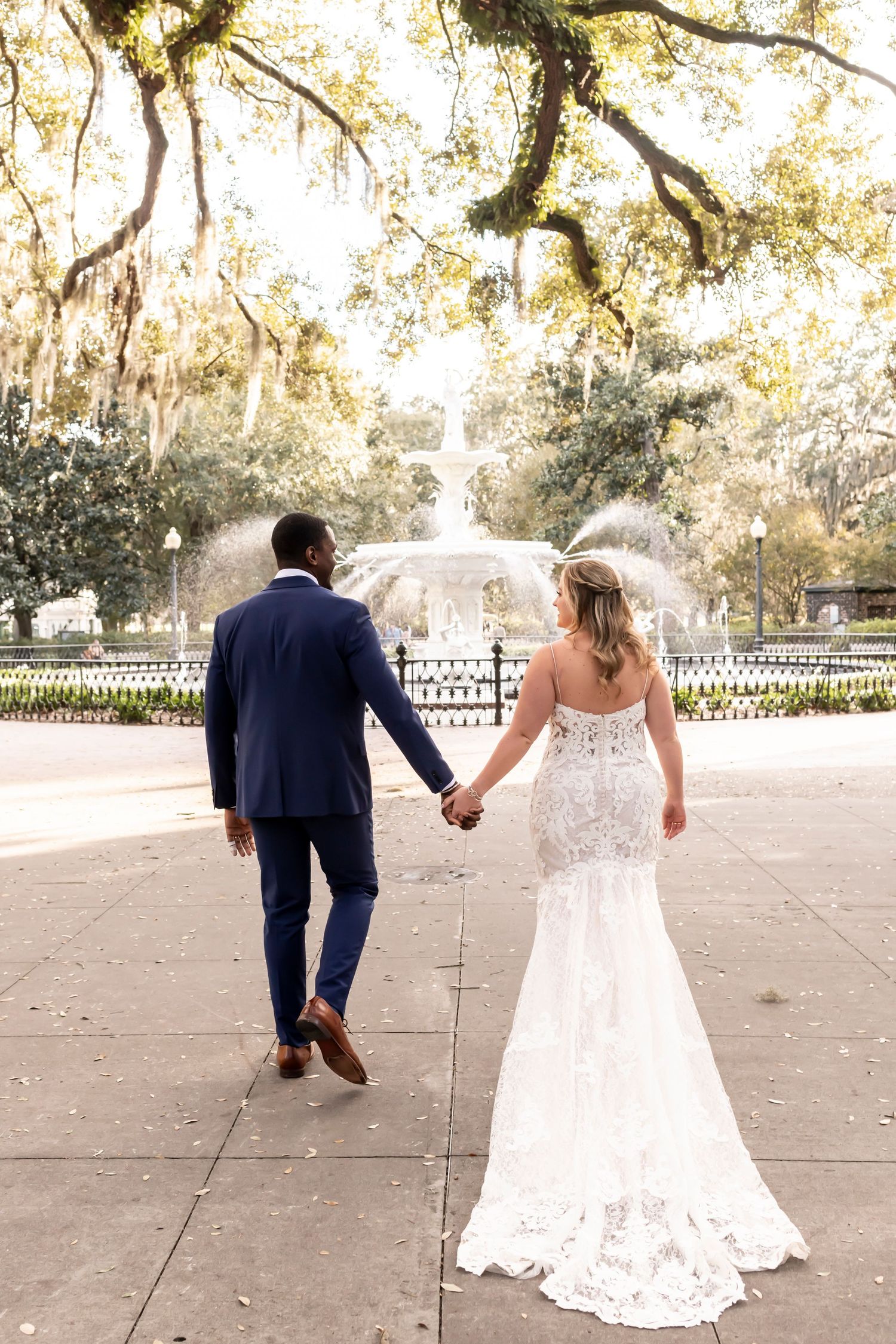 Newlywed couple holding hands while walking toward ornate fountain in garden setting with Spanish moss draped trees.