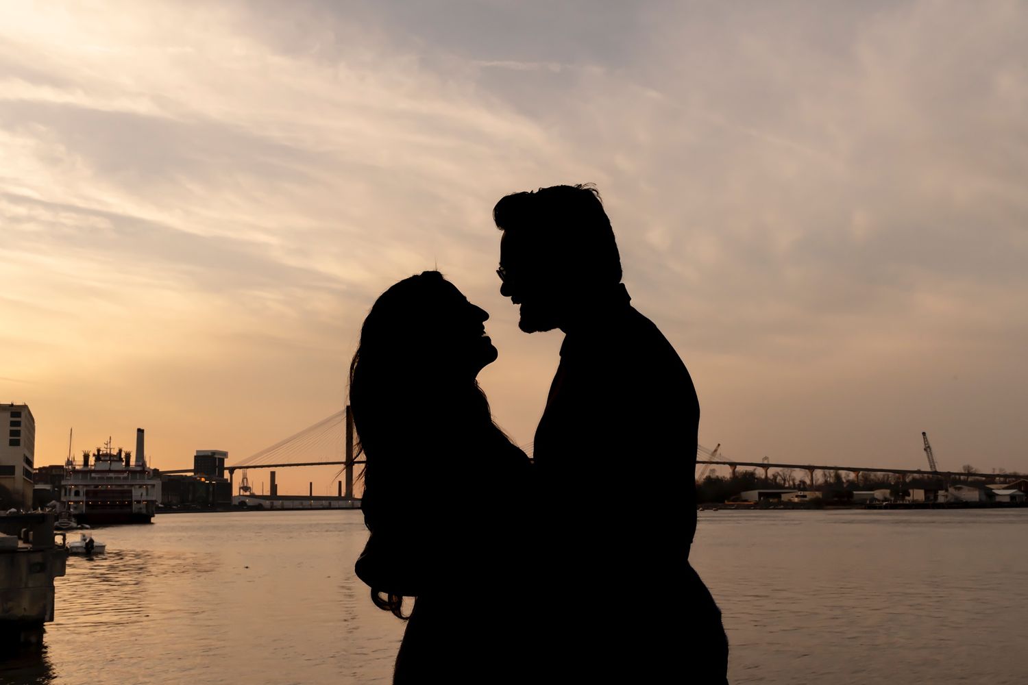 Two silhouetted figures stand together against a sunset sky by the waterfront as a bridge spans the horizon in the distance.