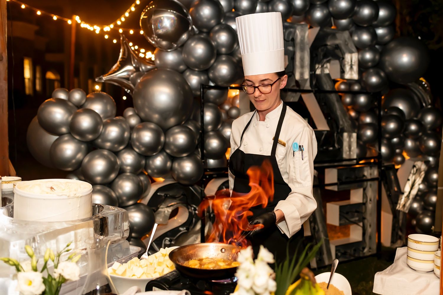 A chef cooking with dramatic flames in front of a decorative black balloon display at an event venue.