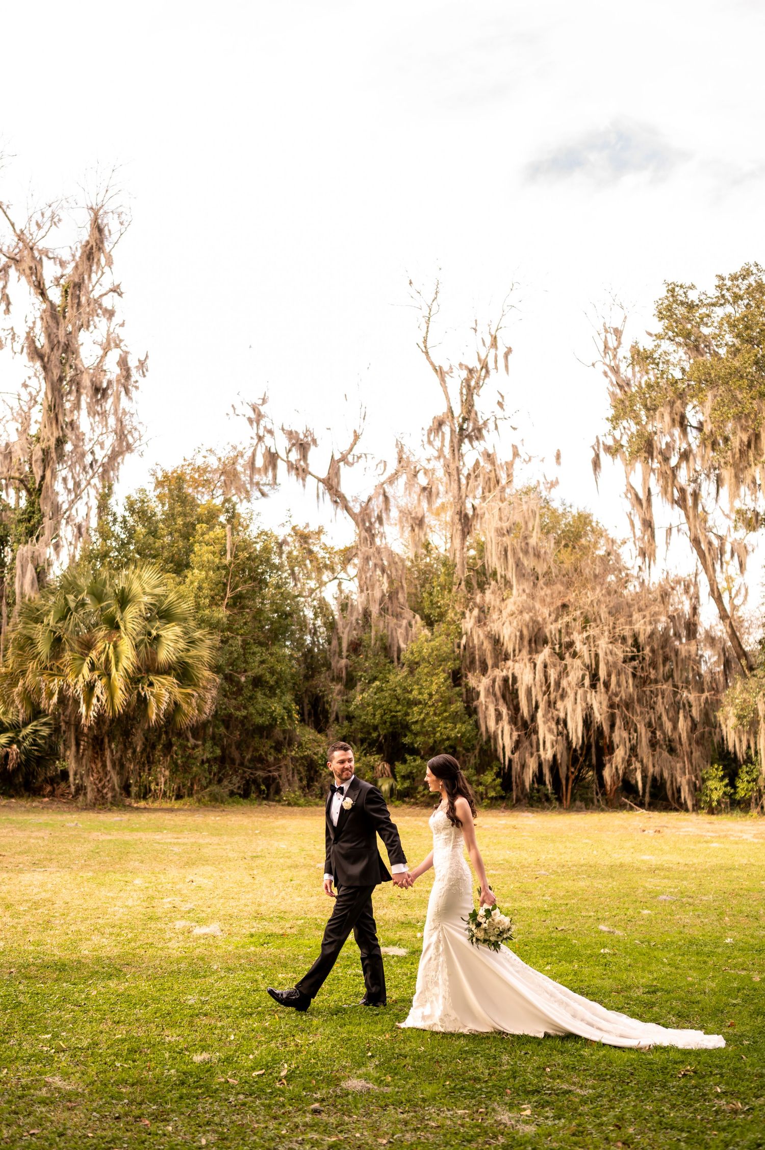 Newlyweds walking on grassy lawn surrounded by Spanish moss draped oak trees in romantic Southern setting.
