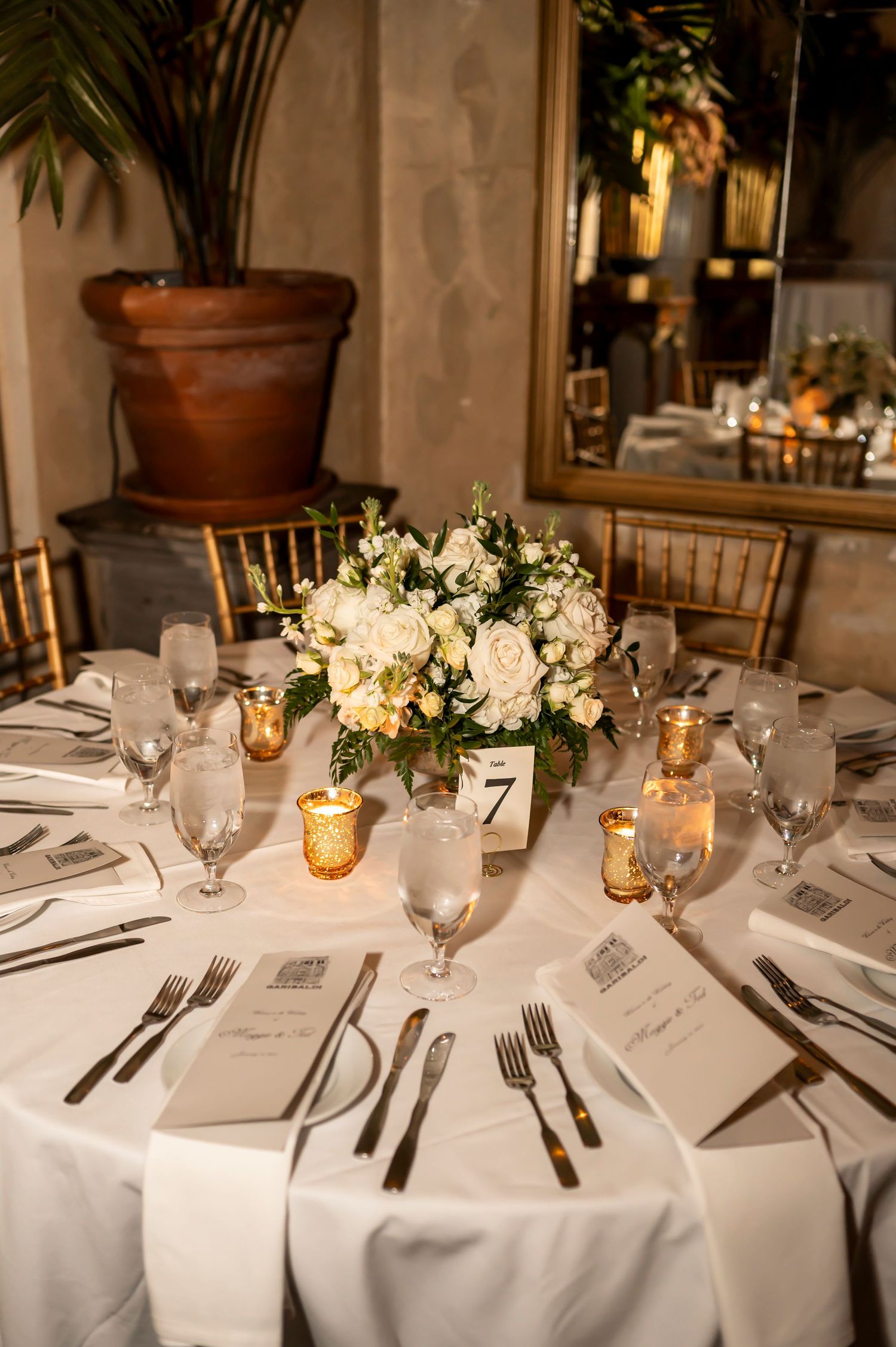 Elegant wedding reception table setting with white floral centerpiece, gold candleholders and place settings at round table.