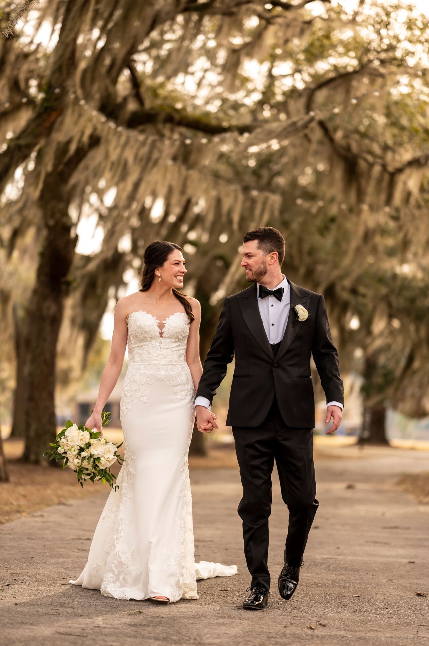 A bride and groom share playful moments while walking along a path lined with Spanish moss draped oak trees.