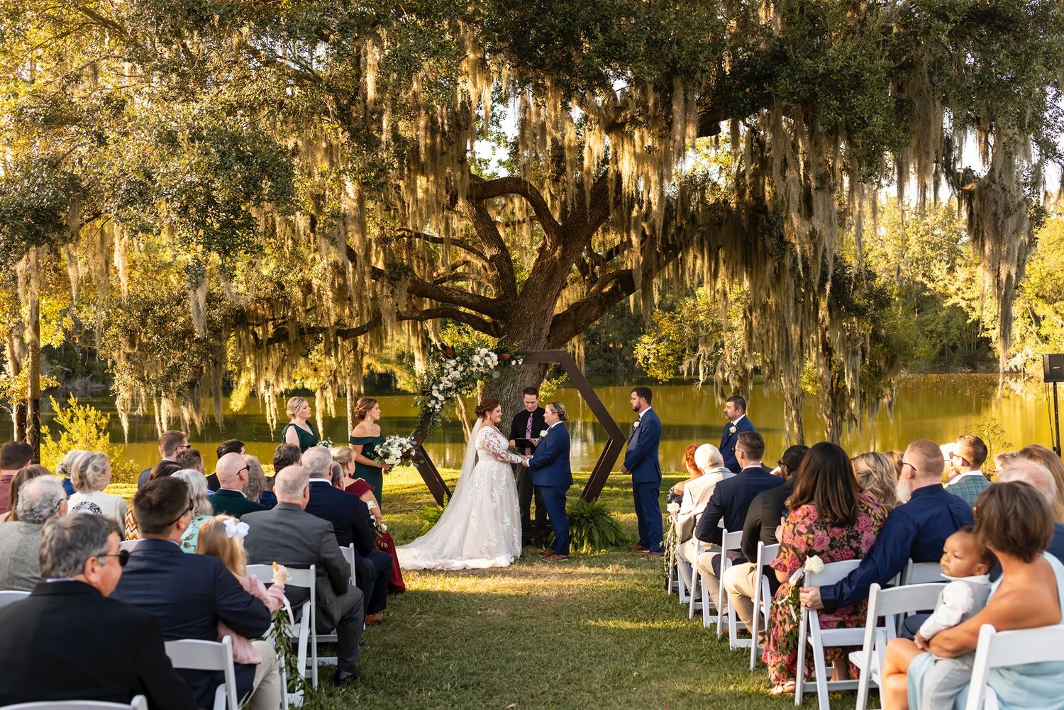 A romantic outdoor wedding ceremony takes place under Spanish moss-draped oak trees at sunset.