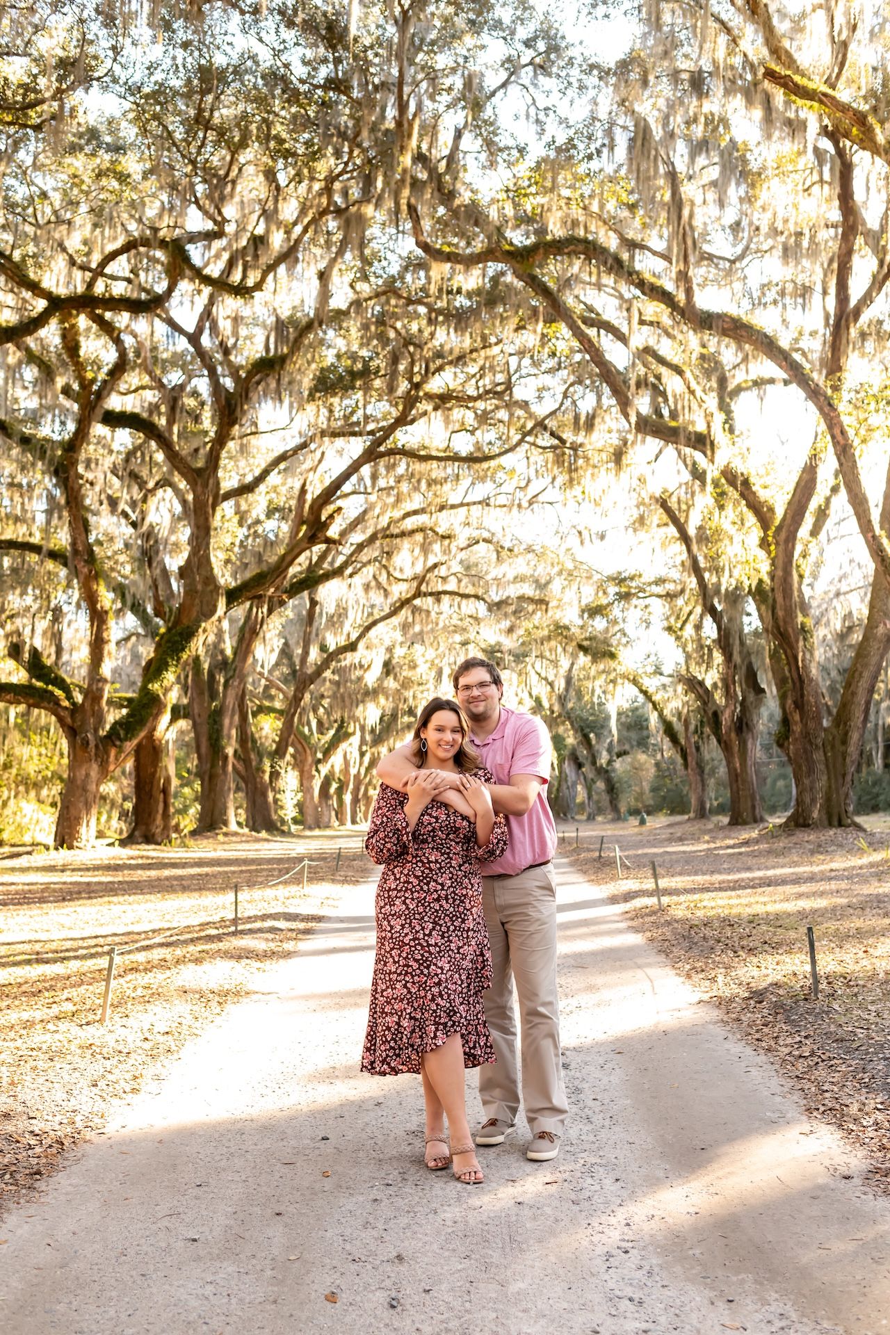 A couple enjoys a romantic stroll under Spanish moss-draped oak trees during golden hour at sunset.
