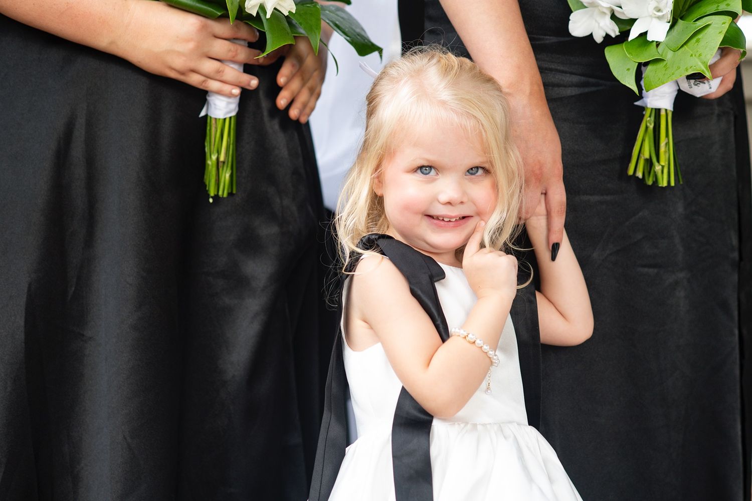 Small child in white dress holding flowers while standing next to adults in formal wear.