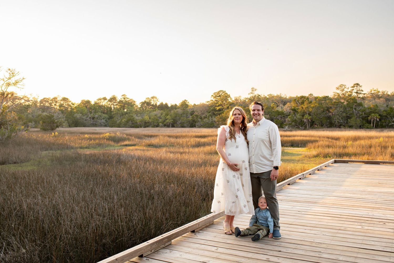 Expecting couple with toddler posing on wooden boardwalk at golden hour over coastal marsh landscape.