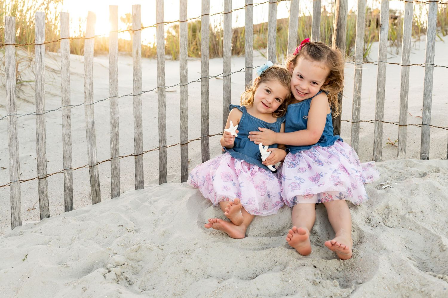 Two children in matching tulle skirts sit together on a sandy beach at sunset with a wooden fence in background.