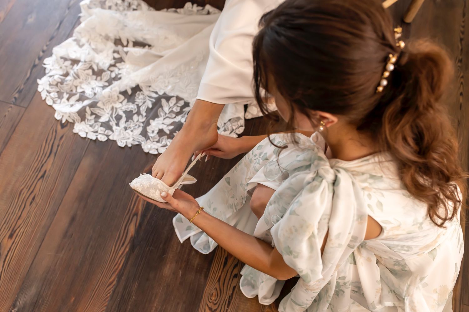 Overhead view of hands adjusting white lace details on wedding dress against wooden floor.