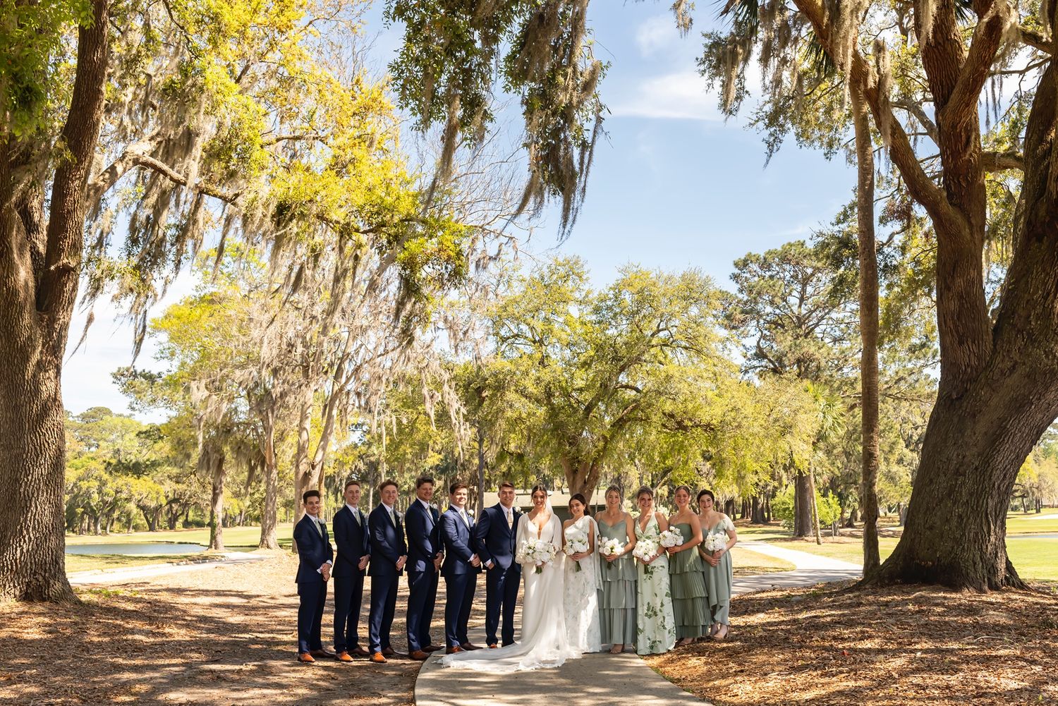 Wedding party stands beneath Spanish moss draped trees on a sunny day in a Southern garden setting.