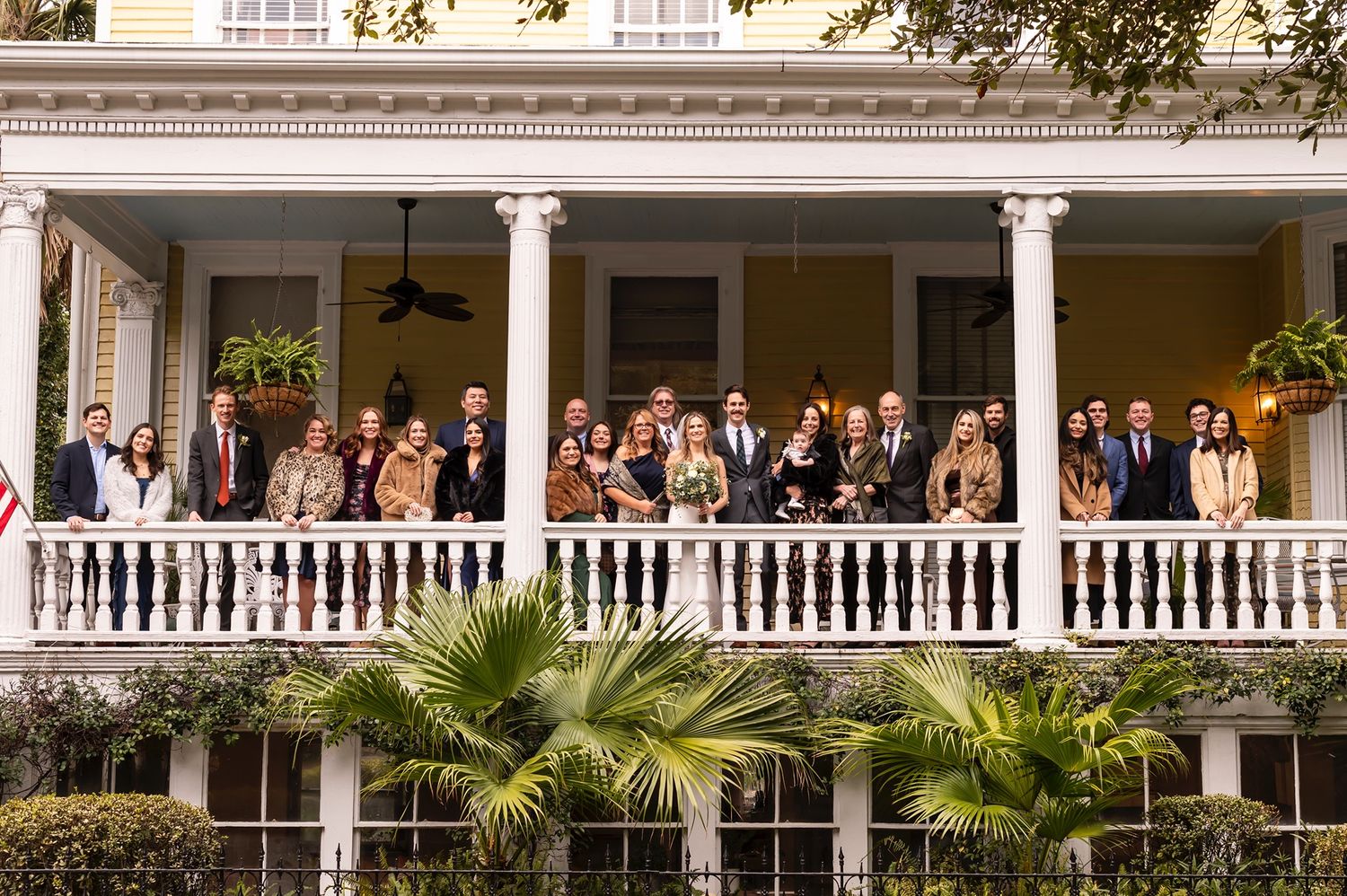 Wedding guests gather on a classic white columned porch decorated with palm trees during an elegant outdoor reception.