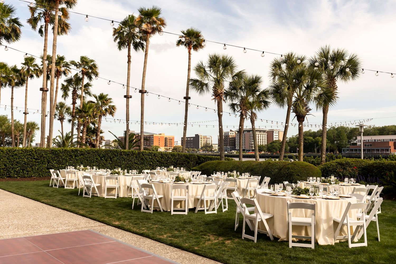 Outdoor wedding reception with white chairs and round tables on green lawn surrounded by tall palm trees with string lights.