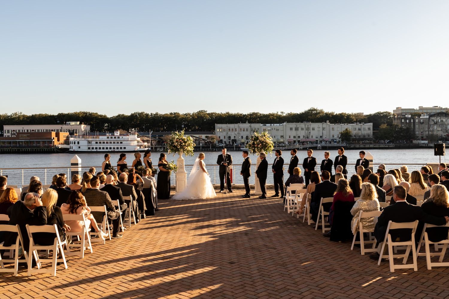 Outdoor waterfront wedding ceremony with bride and groom at altar, guests seated in white chairs on brick plaza at sunset.