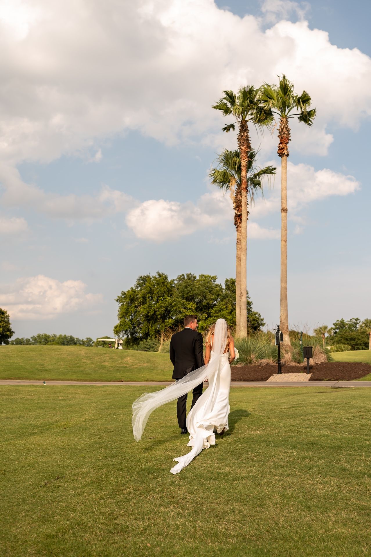 Bride and groom walking on lush green golf course with tall palm trees and cloudy blue sky backdrop.