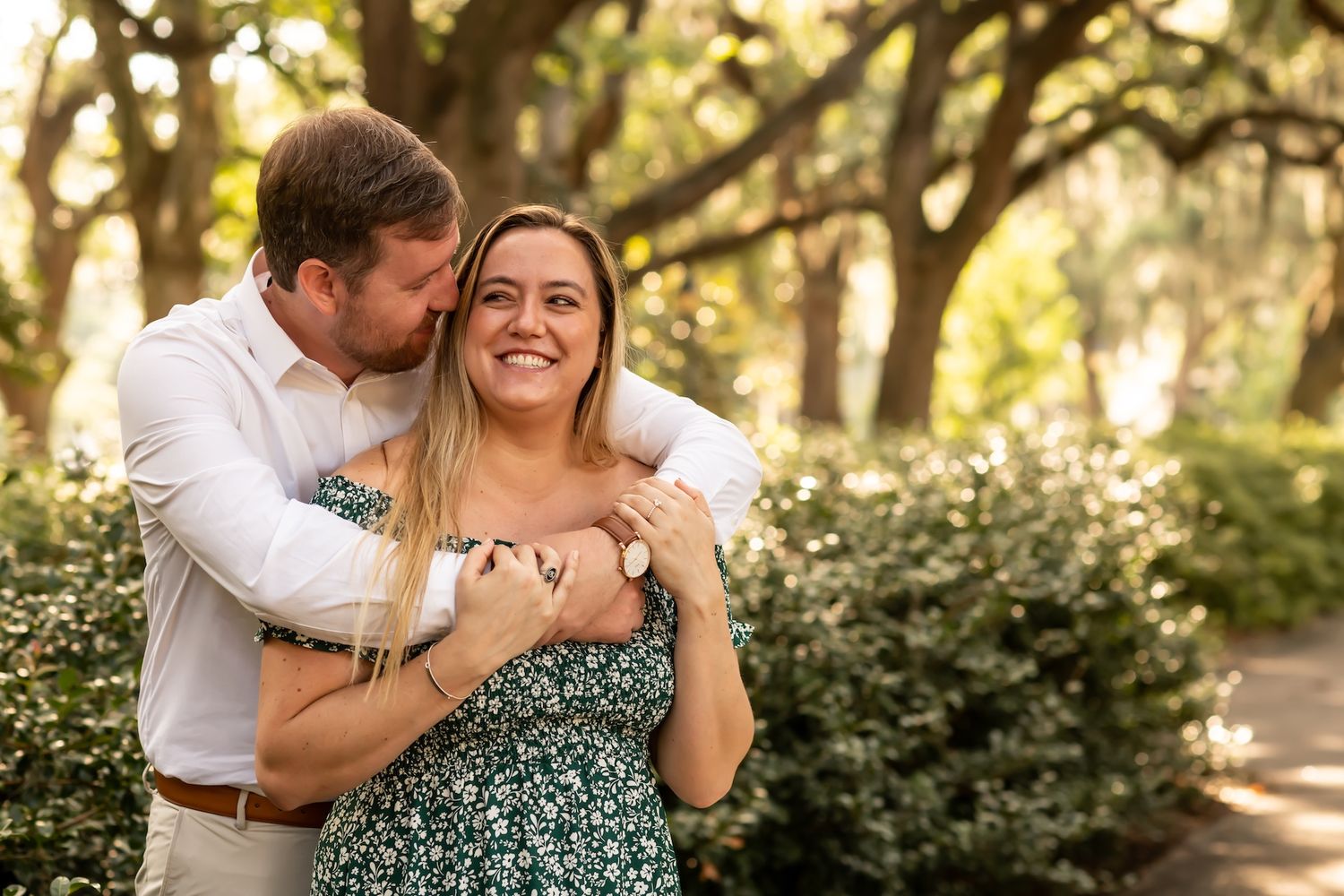 A couple embraces and laughs together on a sunlit garden path with trees and greenery in the background at Forsyth Park.