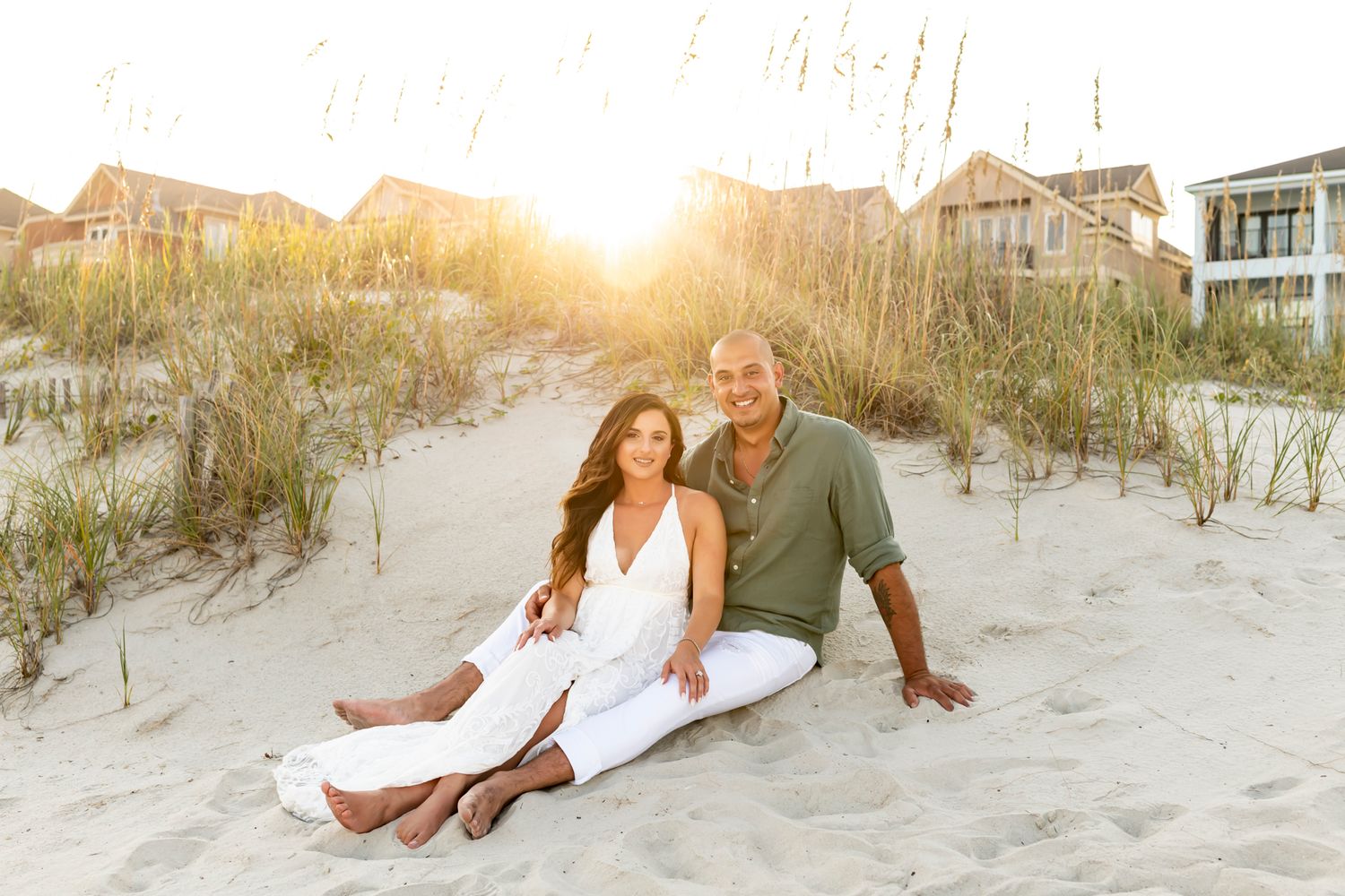 Couple in white clothing posing romantically on a sandy beach at sunset with dunes in the background.
