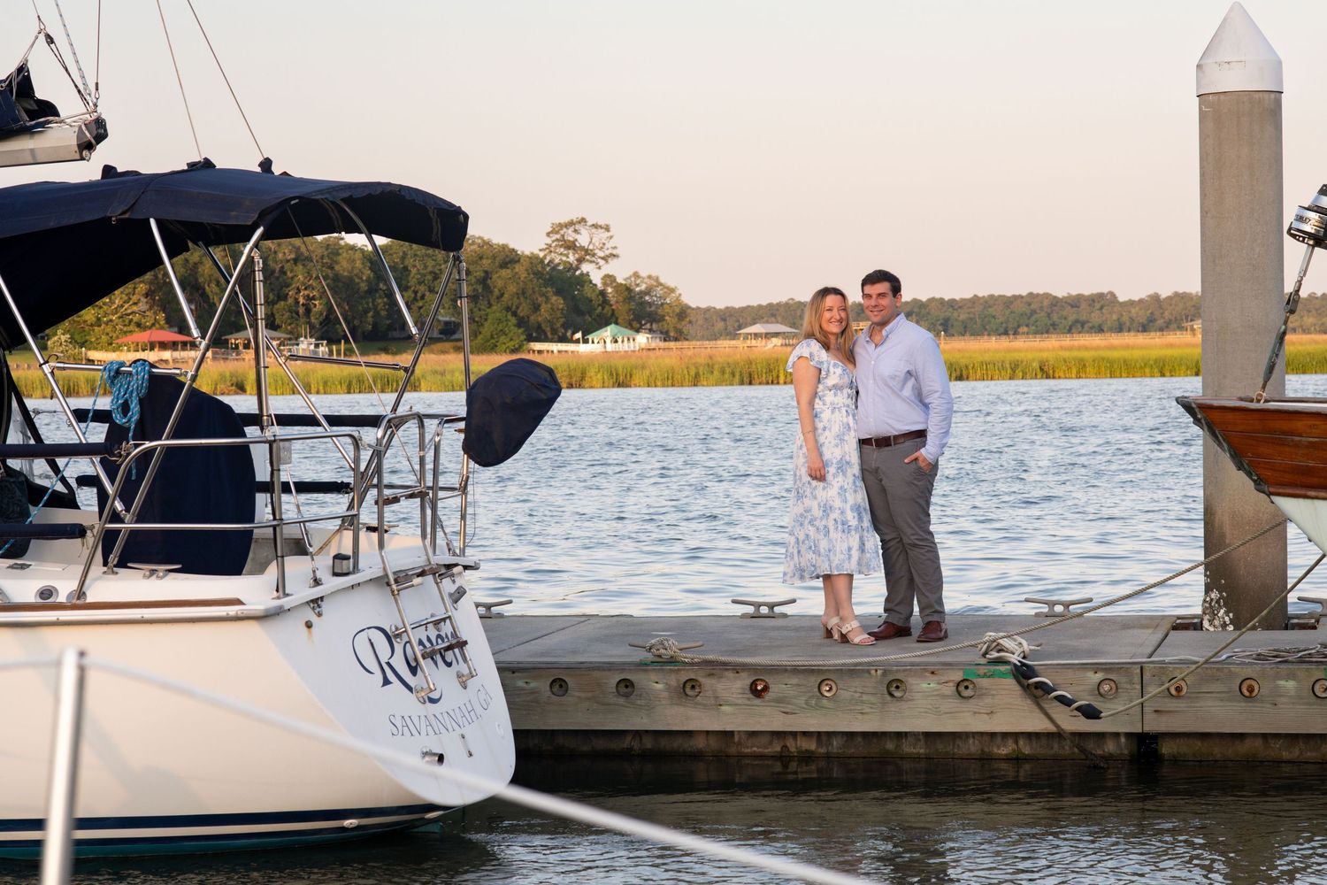 A couple stands together on a wooden dock at sunset next to a sailboat in a coastal marina setting.