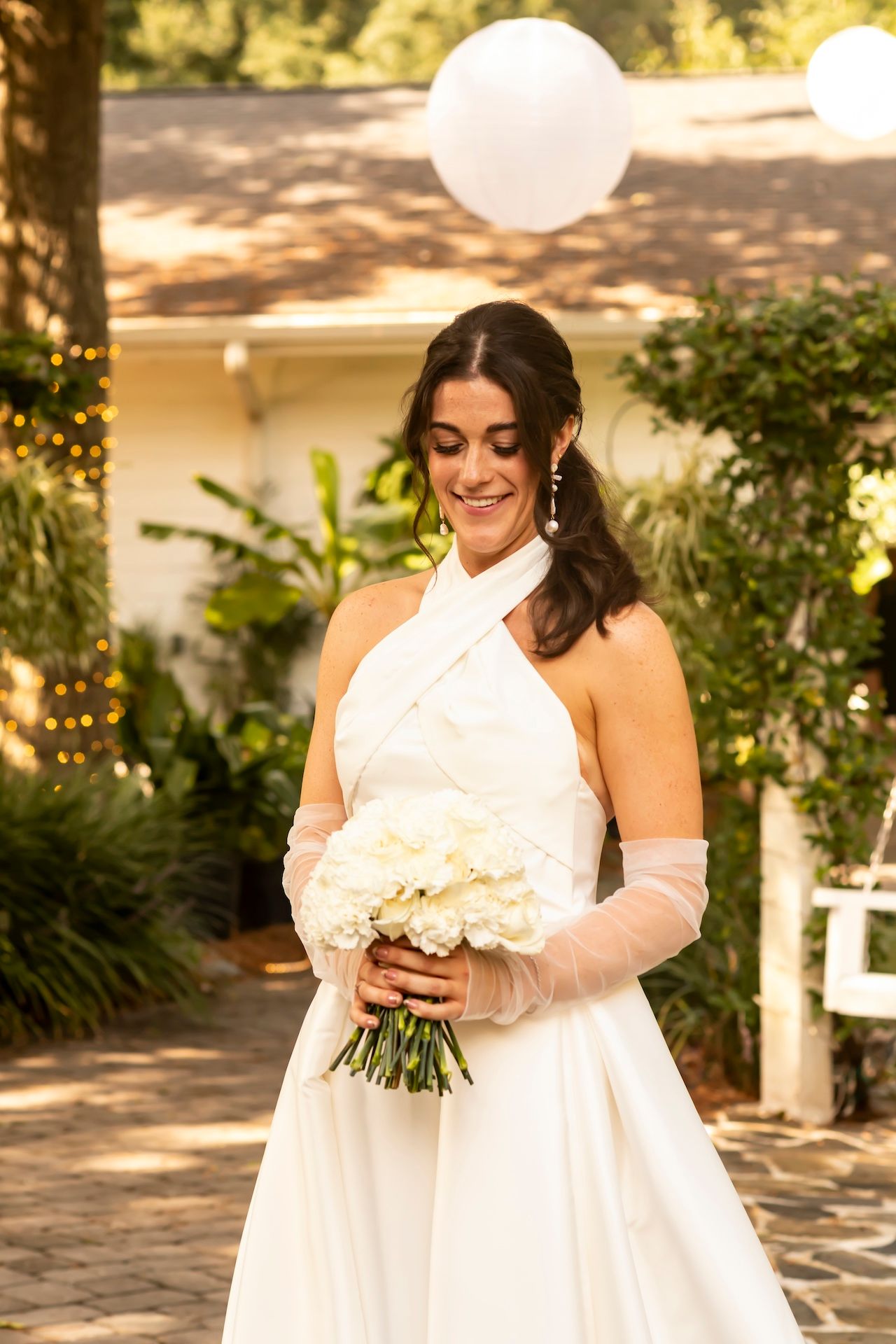 Bride in white halter dress and gloves holding a bouquet of white flowers in a garden setting with string lights.