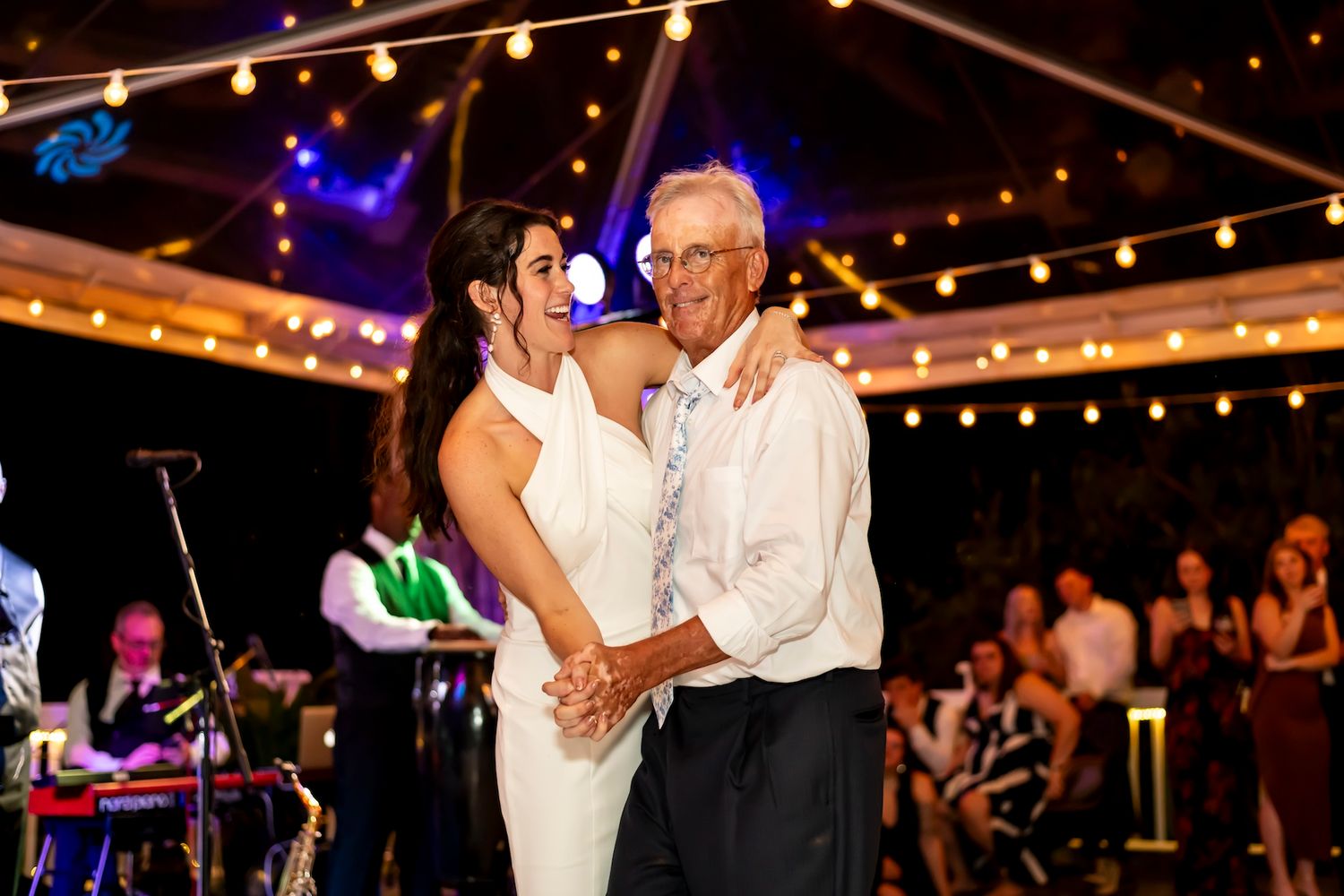 A couple shares a joyful dance together at an evening reception under string lights with a band performing in the background.
