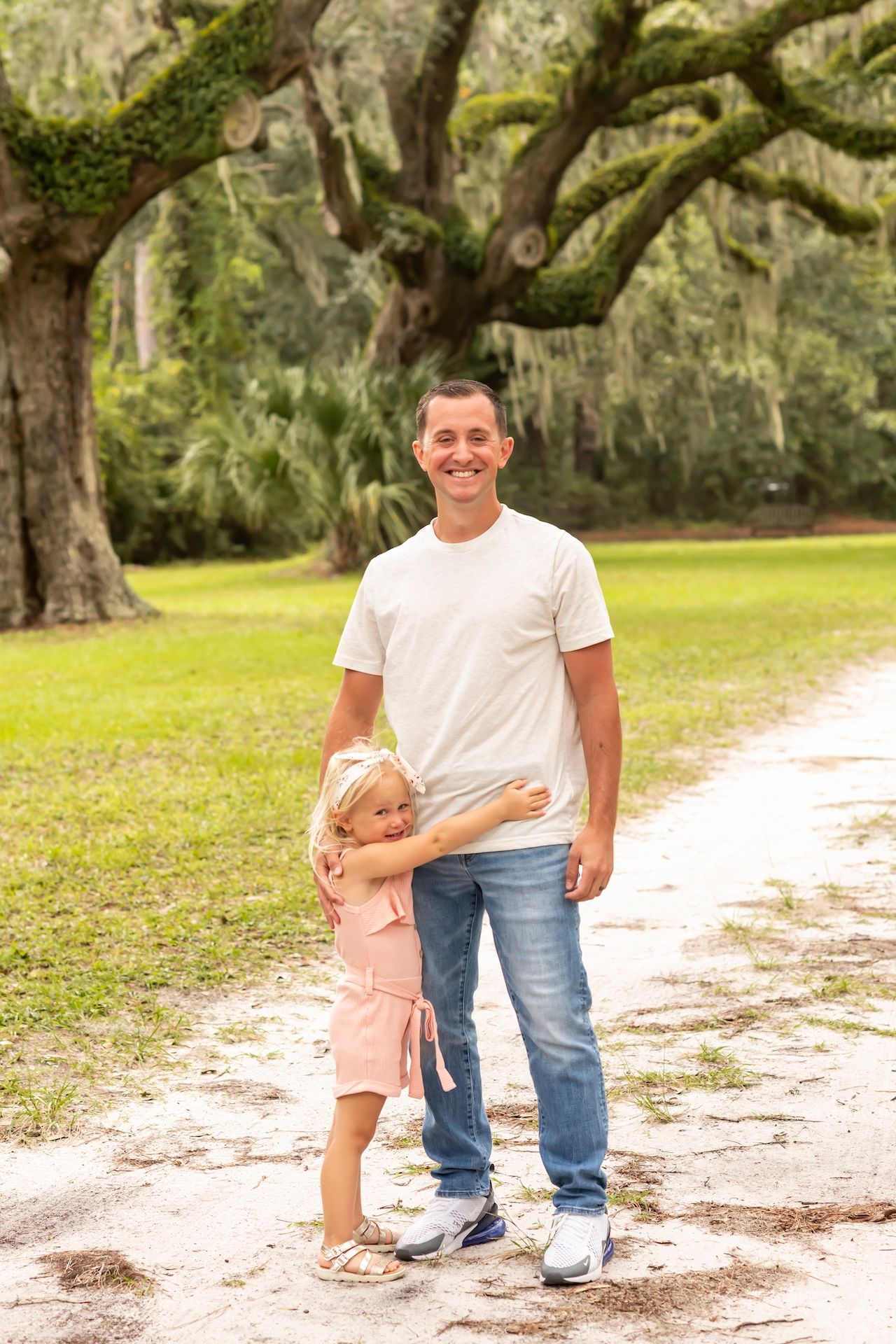 A person in a white t-shirt and jeans stands with a small child on a tree-lined dirt path in a sunny park.