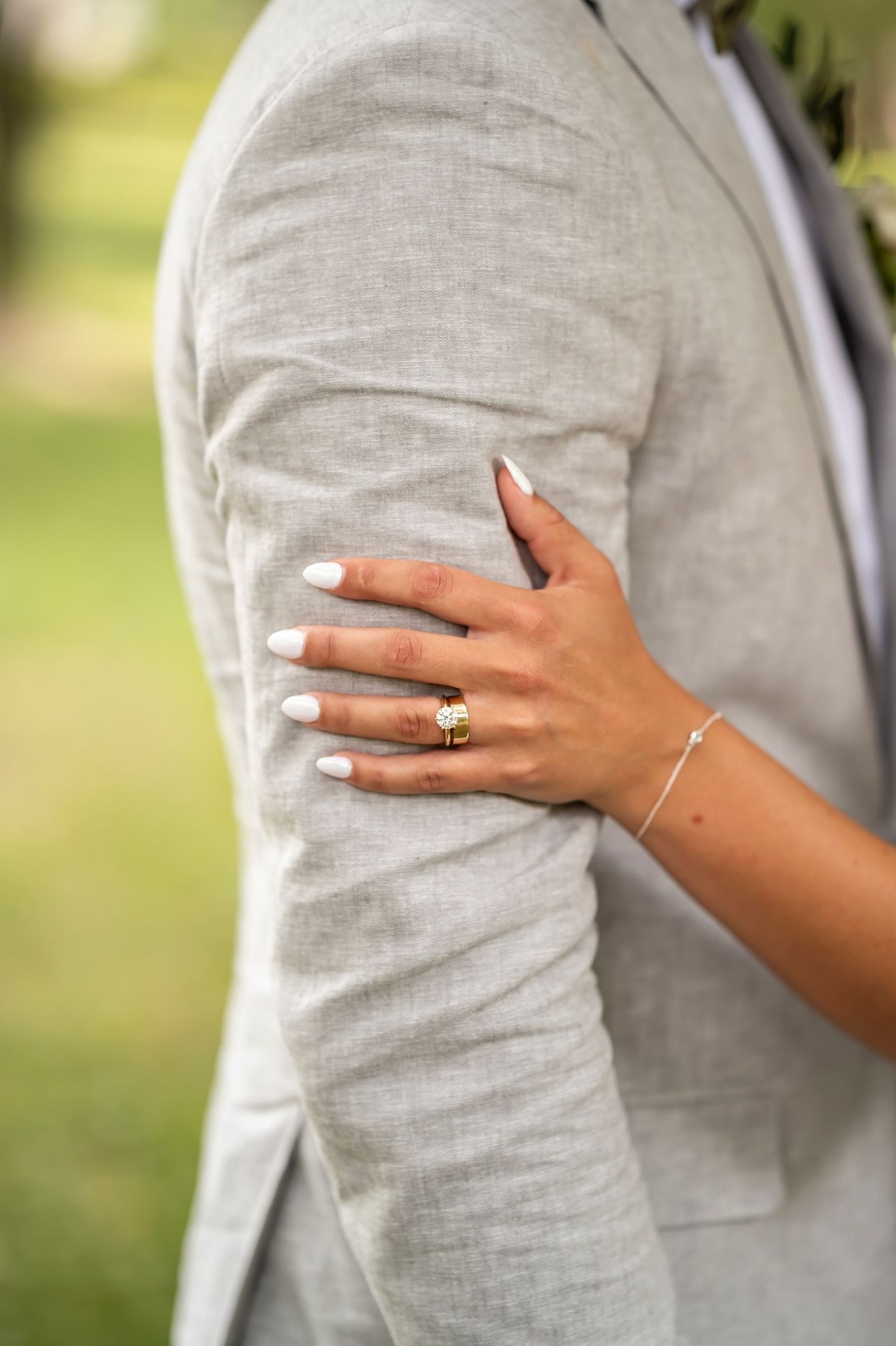 A hand with white nail polish and gold jewelry rests on a light gray suit sleeve in an outdoor setting.
