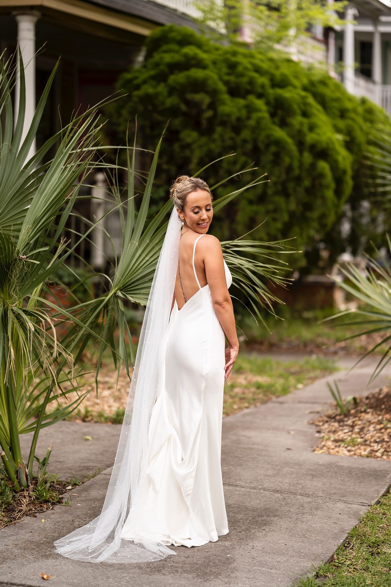 Elegant white wedding dress with low back and flowing train on walkway surrounded by tropical palm fronds.