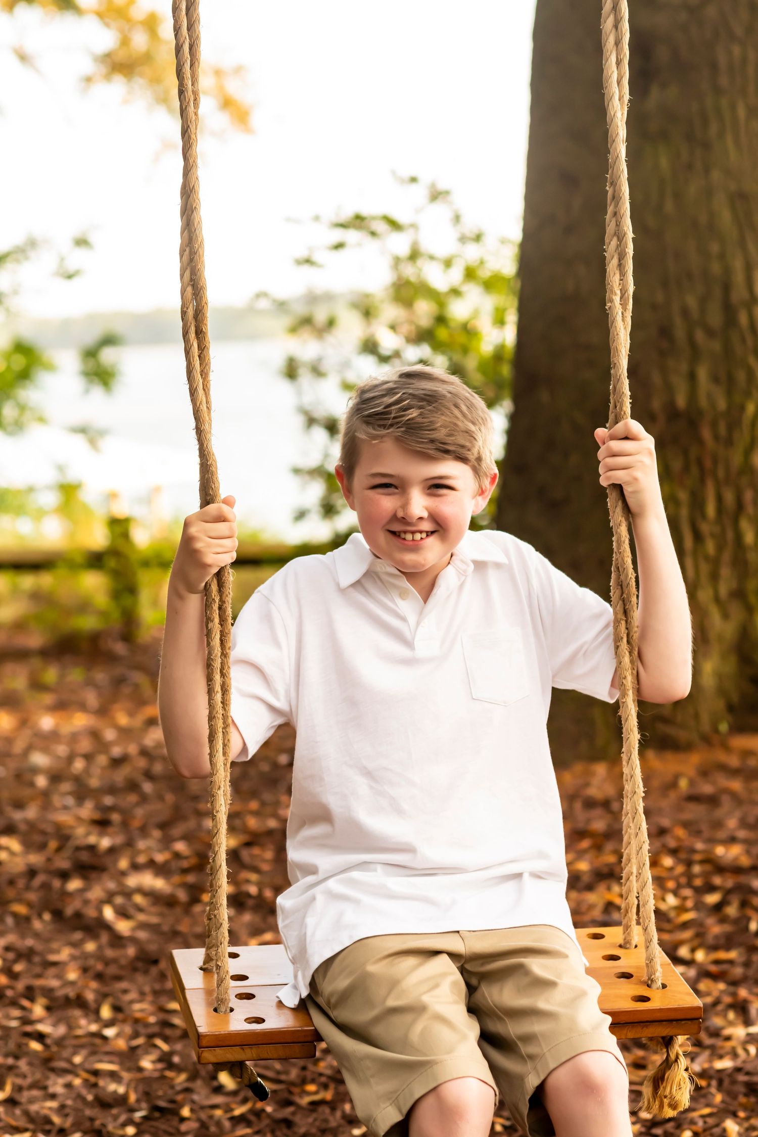 A young person in a white shirt and khaki shorts sits happily on a wooden rope swing in a wooded outdoor setting.