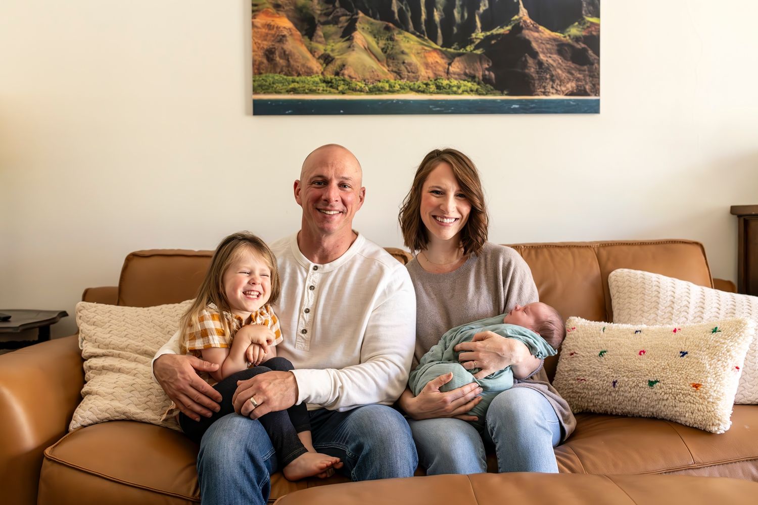 Family sitting together on brown leather couch with newborn baby and toddler in cozy living room.