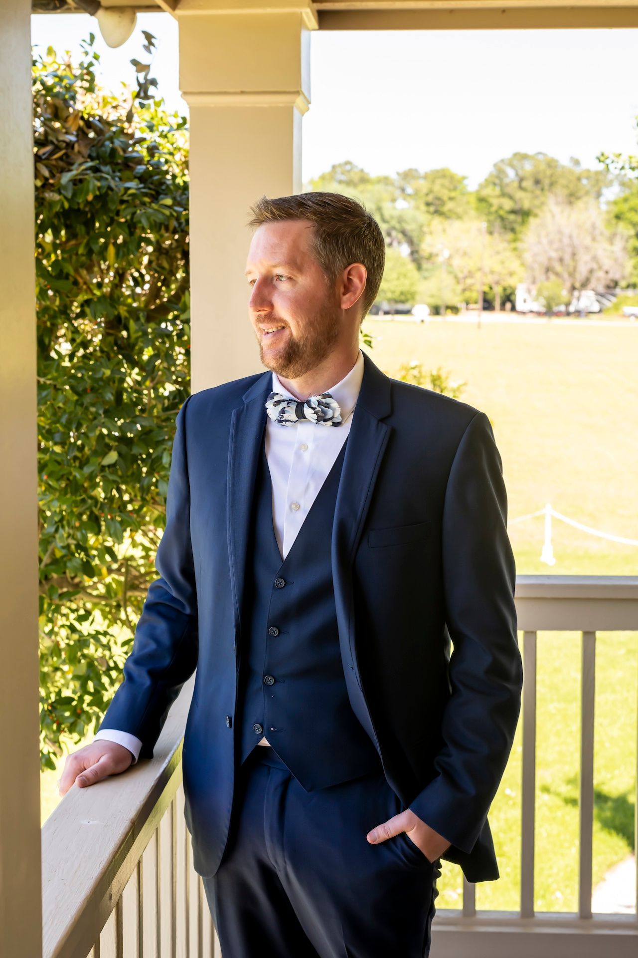 A well-dressed person in a navy three-piece suit and bow tie stands on a porch looking off into the distance.