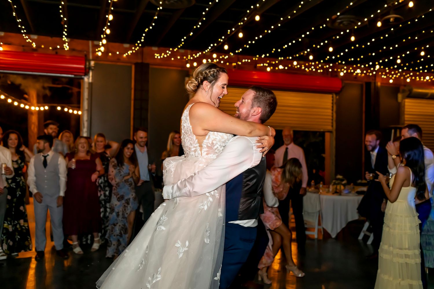 A couple shares their first dance under glowing string lights at their wedding reception while guests look on.