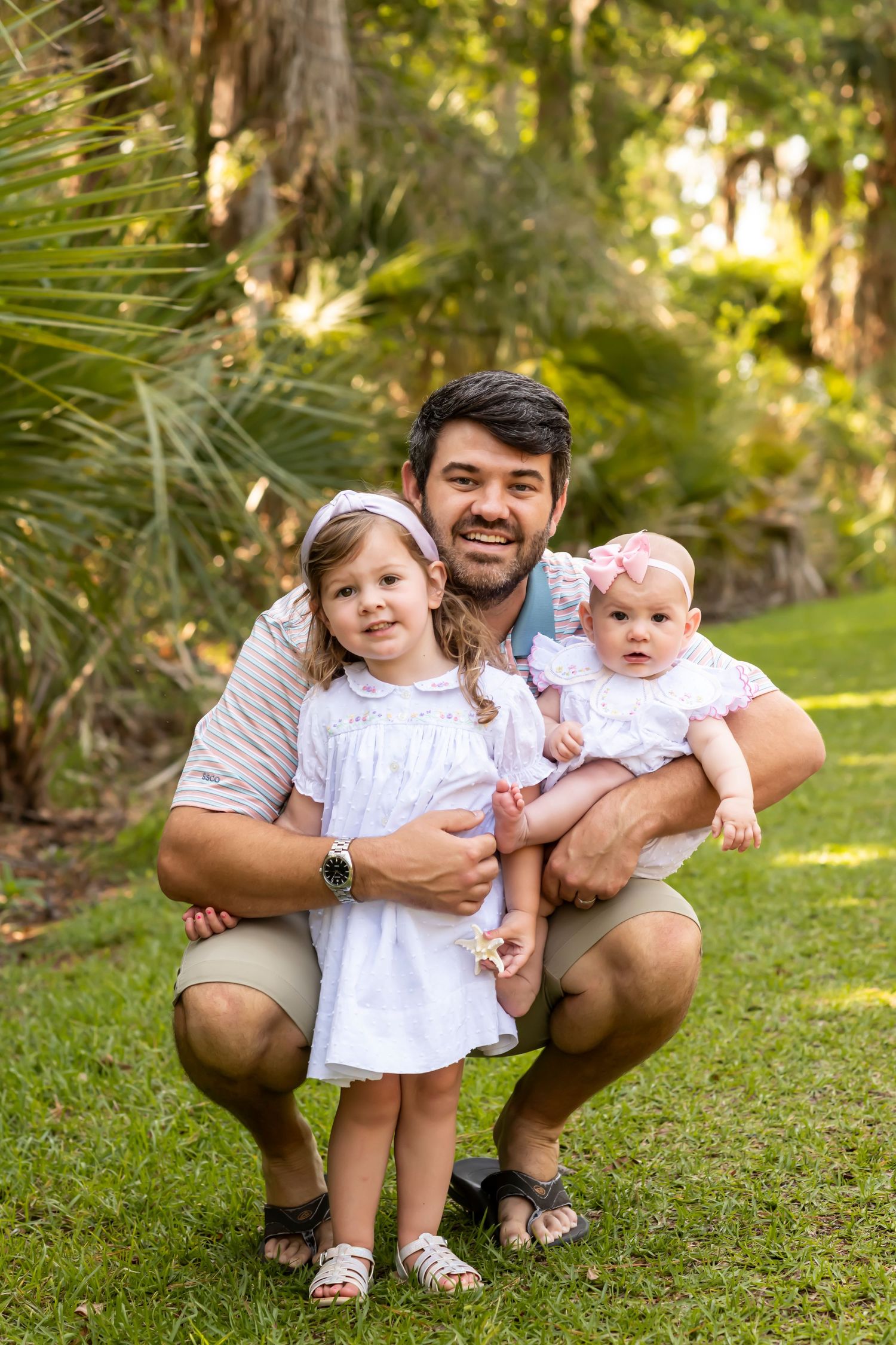 Group crouching together on green grass in a sunny wooded park setting wearing white and khaki colors.