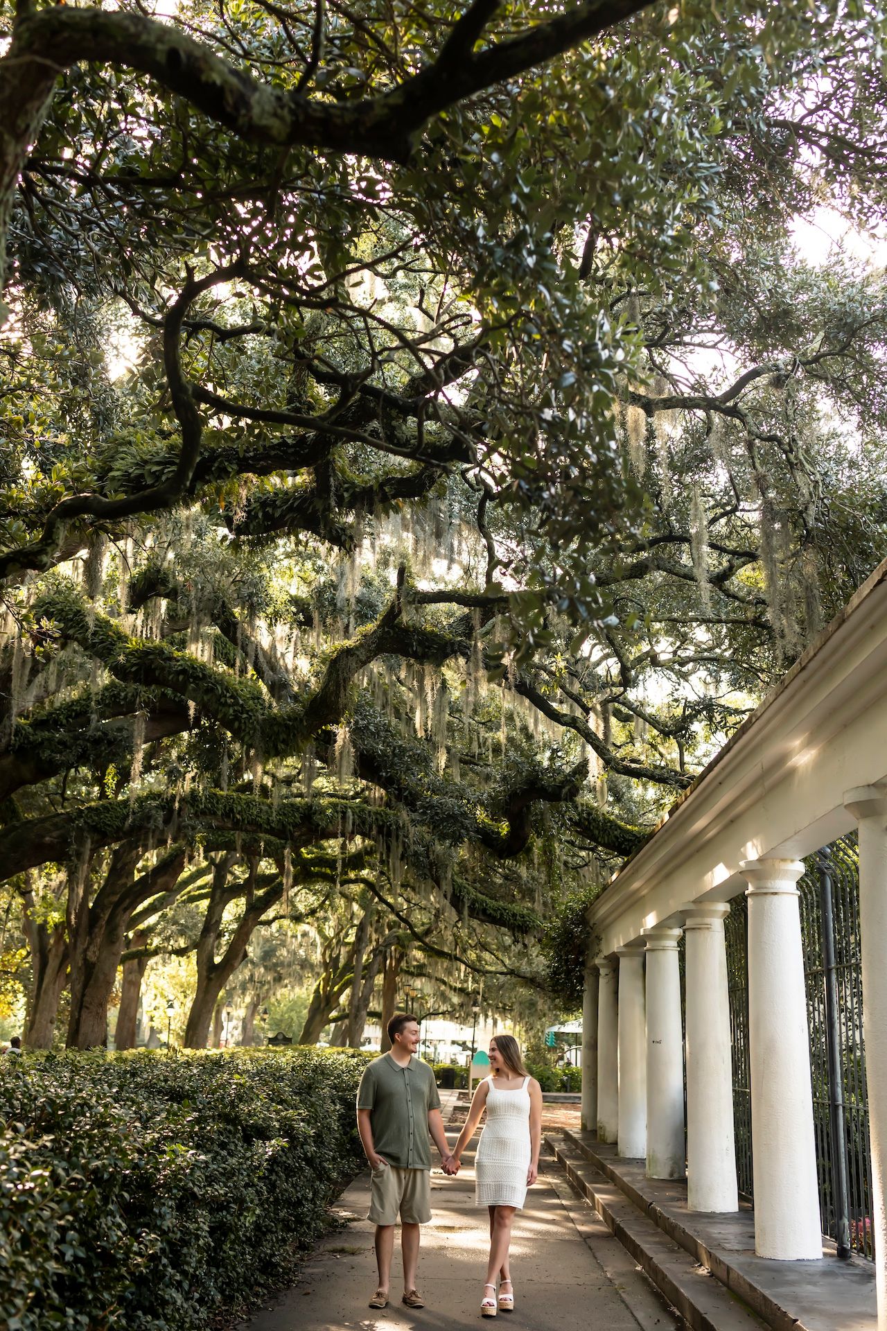 A couple walks beneath majestic Spanish moss-draped oak trees along a columned walkway in historic Savannah, Georgia.